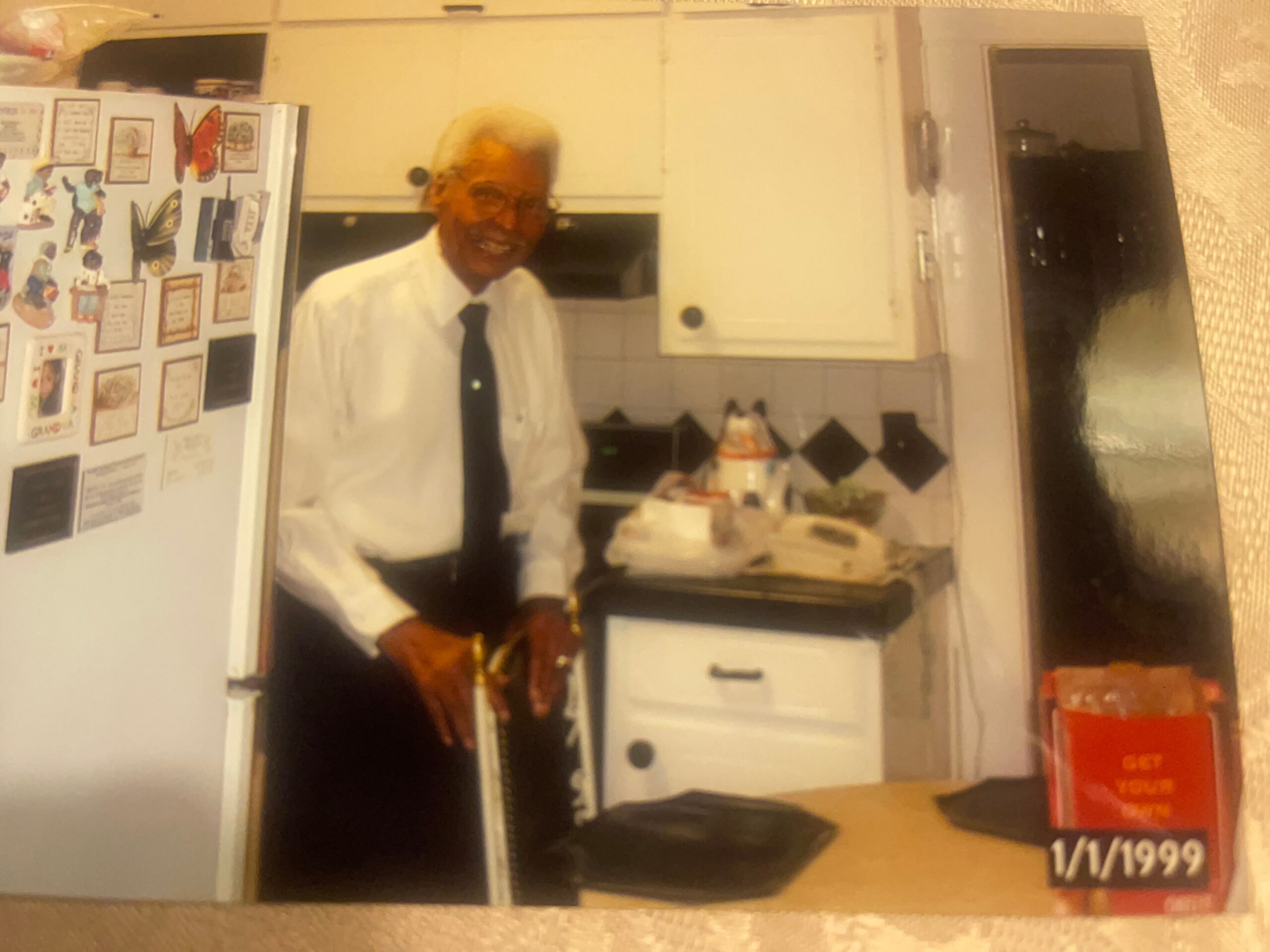 Older man in a white shirt and black tie standing in a kitchen by a refrigerator, smiling at the camera beside a cake on the counter.