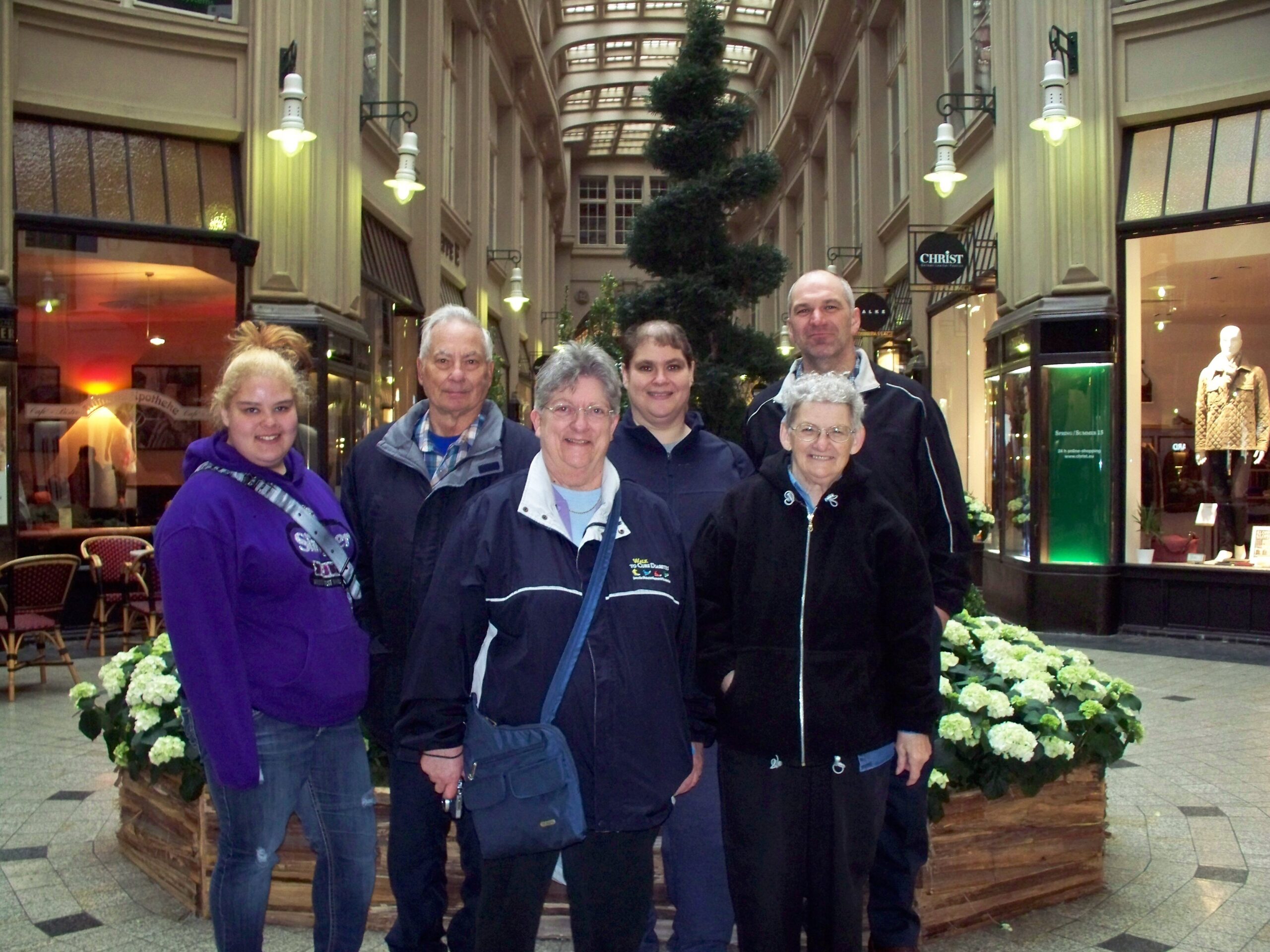 Five people posing for a group photo in an indoor shopping arcade, with a tall spiral evergreen plant behind them behind a circular planter.