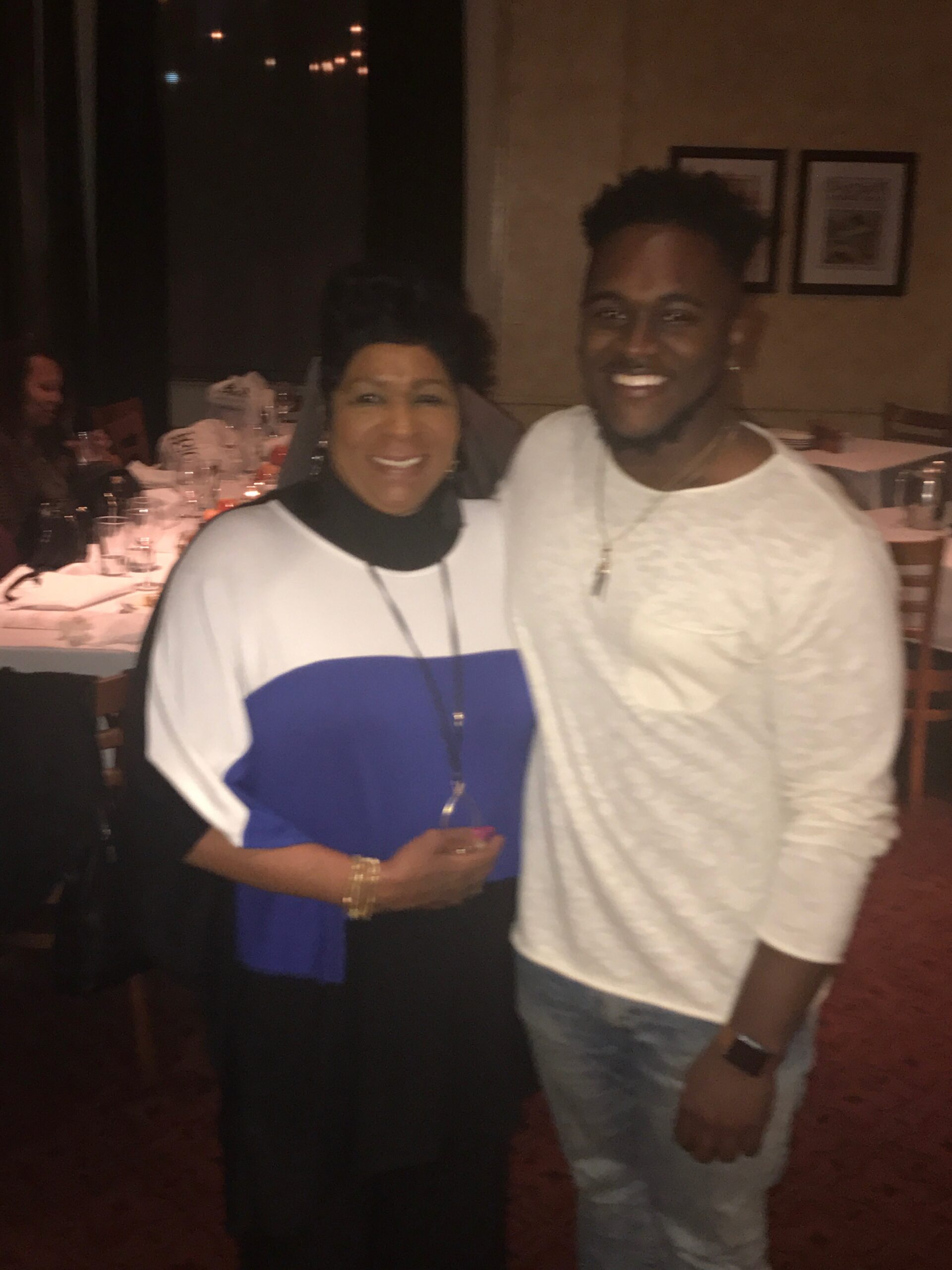 Two friends posing and smiling at a dim banquet hall, woman in white top with blue panel and man in white long-sleeve shirt standing together near a candlelit table.