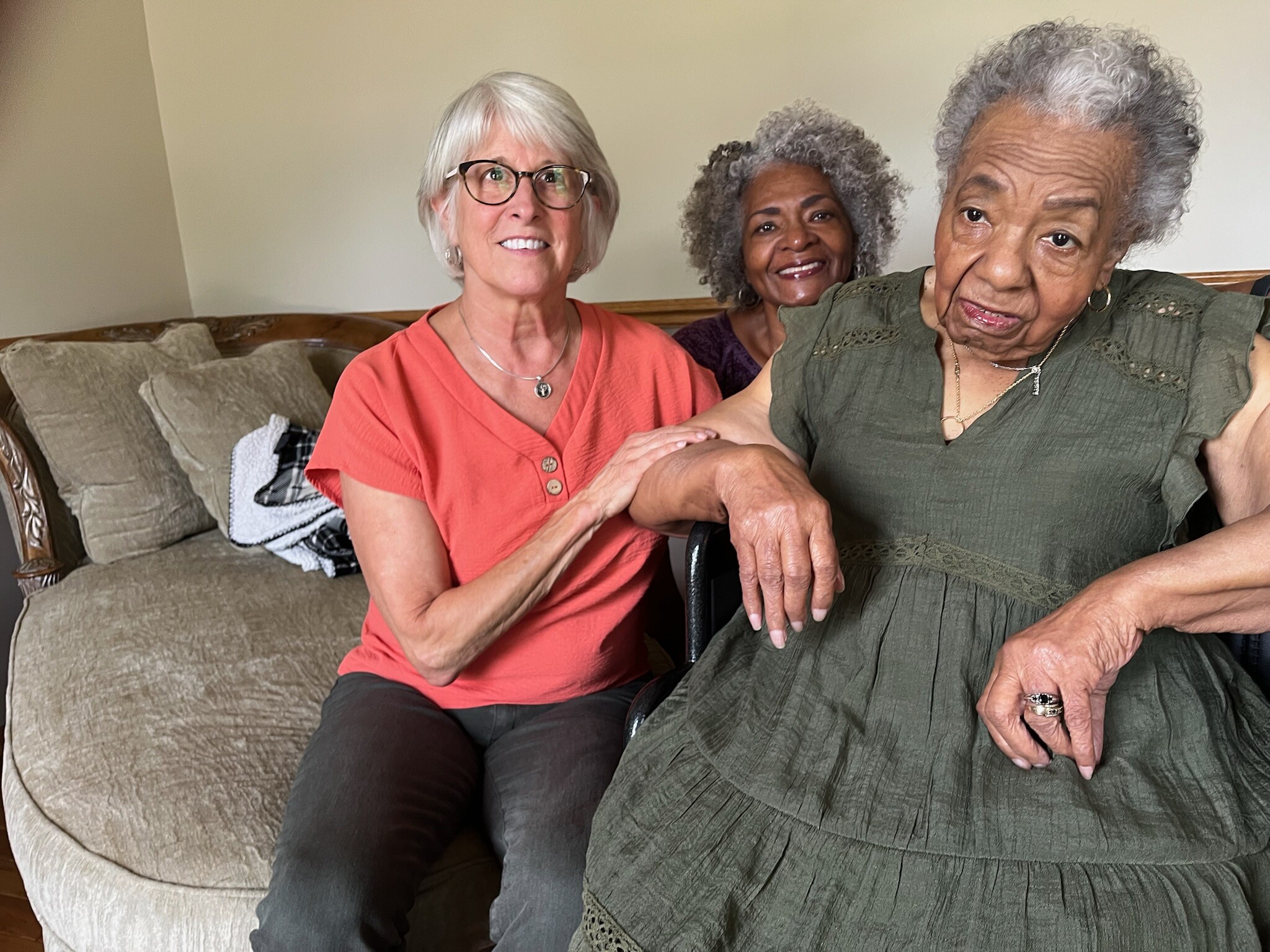 Three smiling elderly women sit close together on a beige sofa, arms around each other, posing for the photo.
