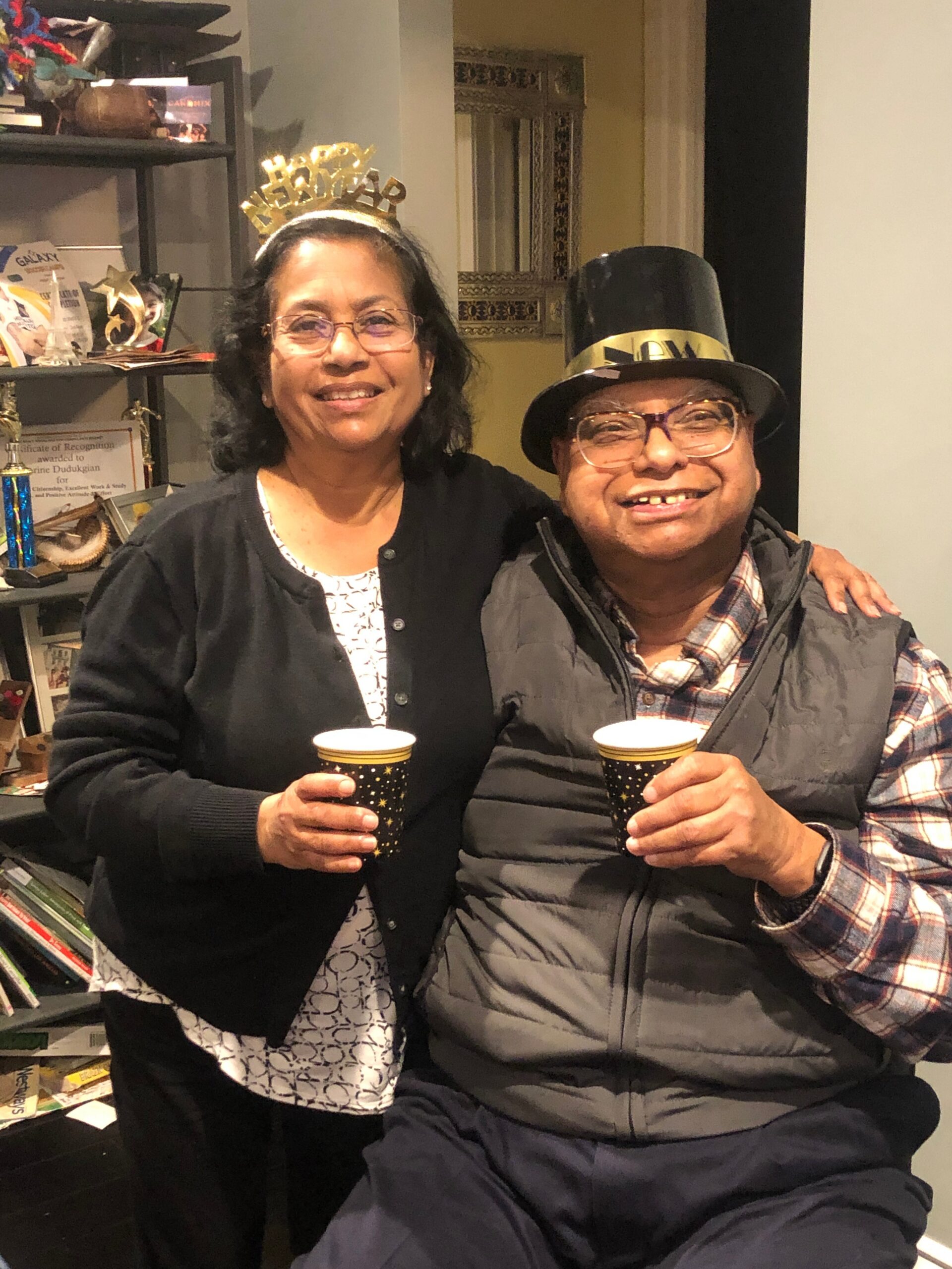 Two smiling people celebrate indoors, each holding a starry paper cup; woman wears a gold 'Happy Birthday' crown and black cardigan, man wears a black top hat with a gold band and a puffy vest.