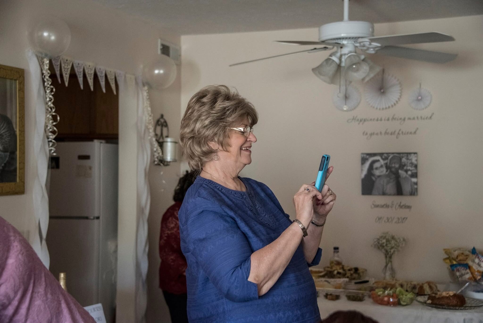 Smiling woman in a blue blouse holding a teal smartphone at a party, with balloons and decorations in the background.