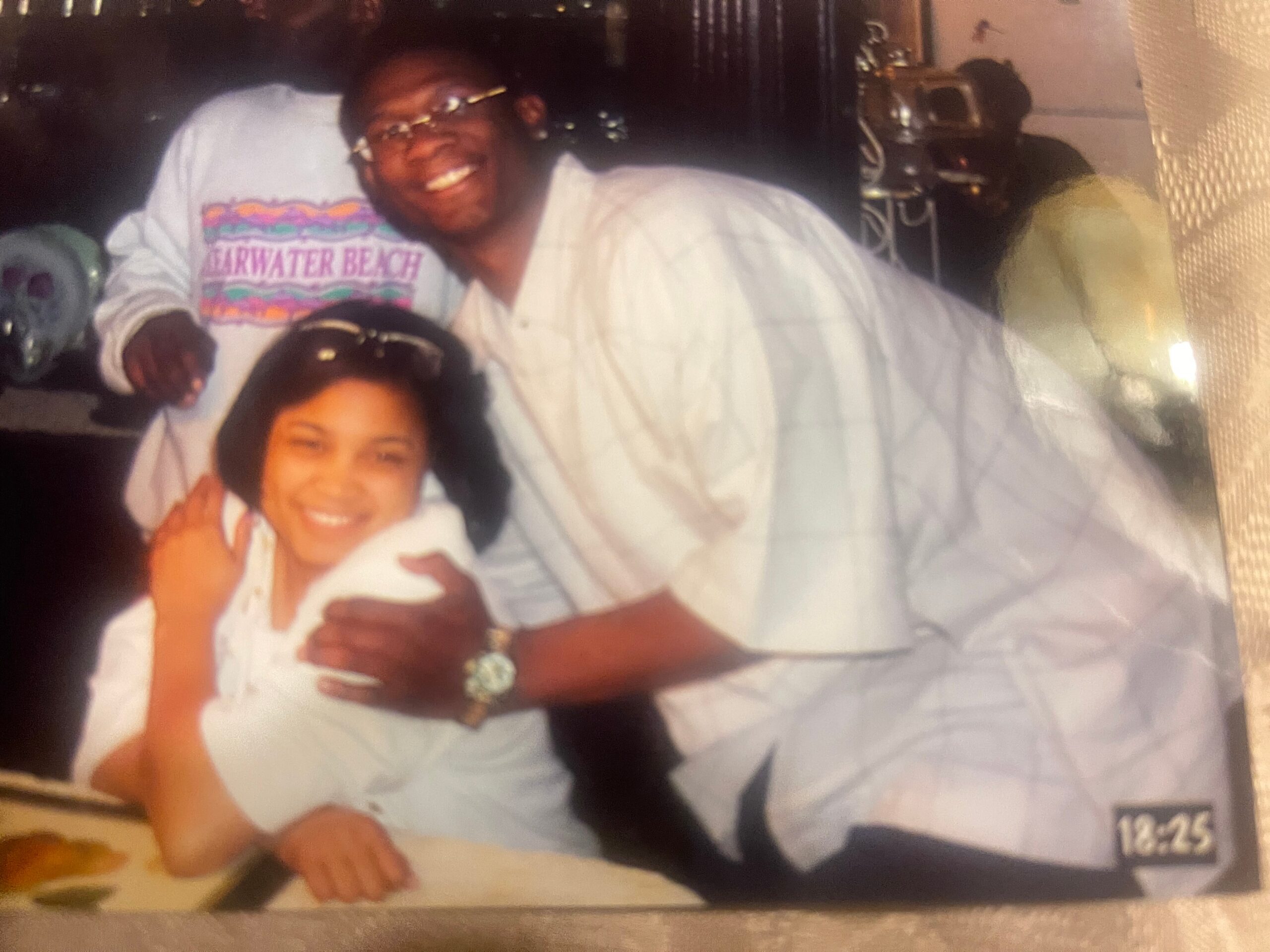 Smiling man with glasses in a white shirt leaning over a table beside a smiling woman indoors.