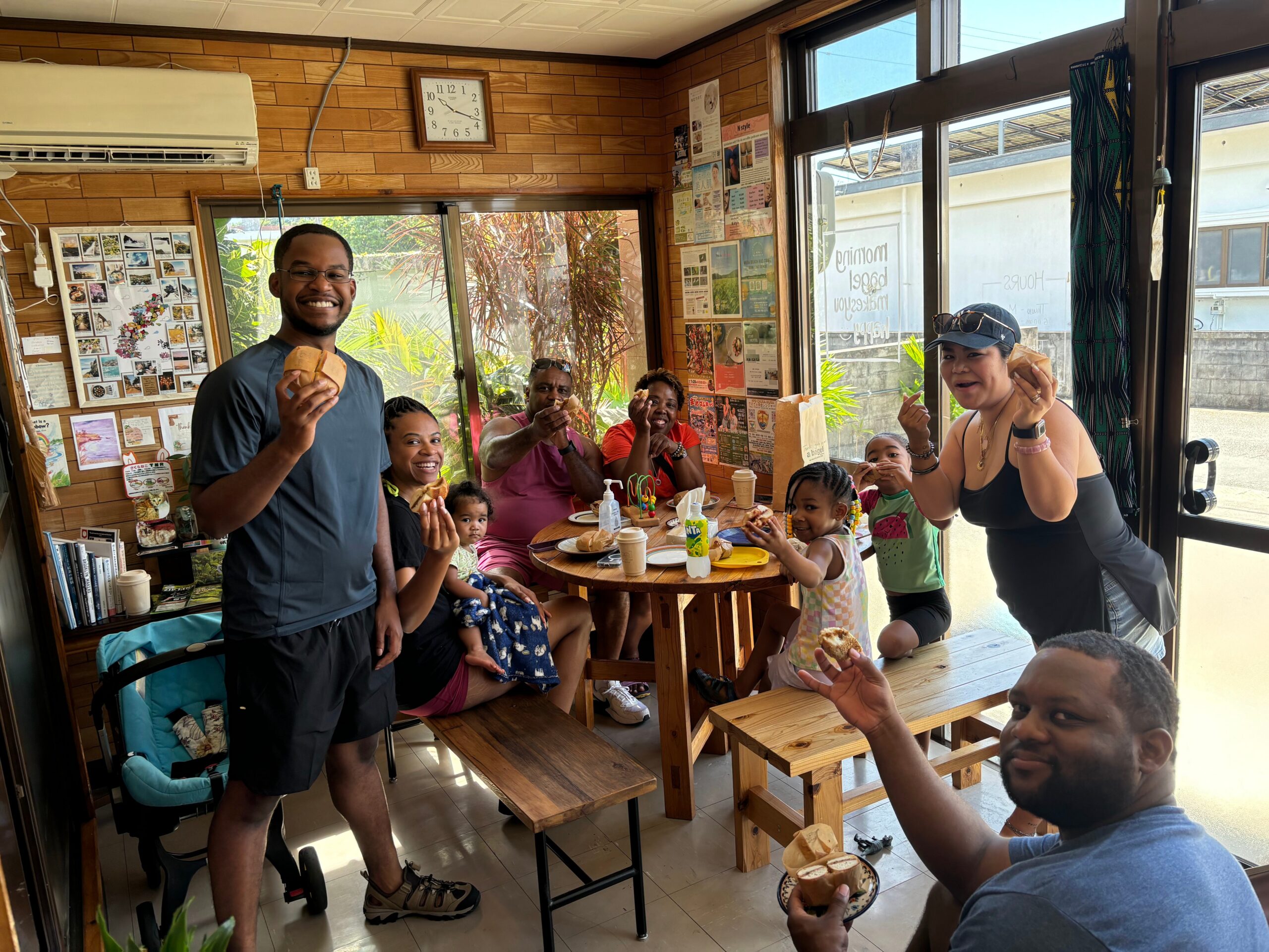 Group of adults and children around a wooden table, smiling and holding bread rolls in a bright, casual cafe/restaurant