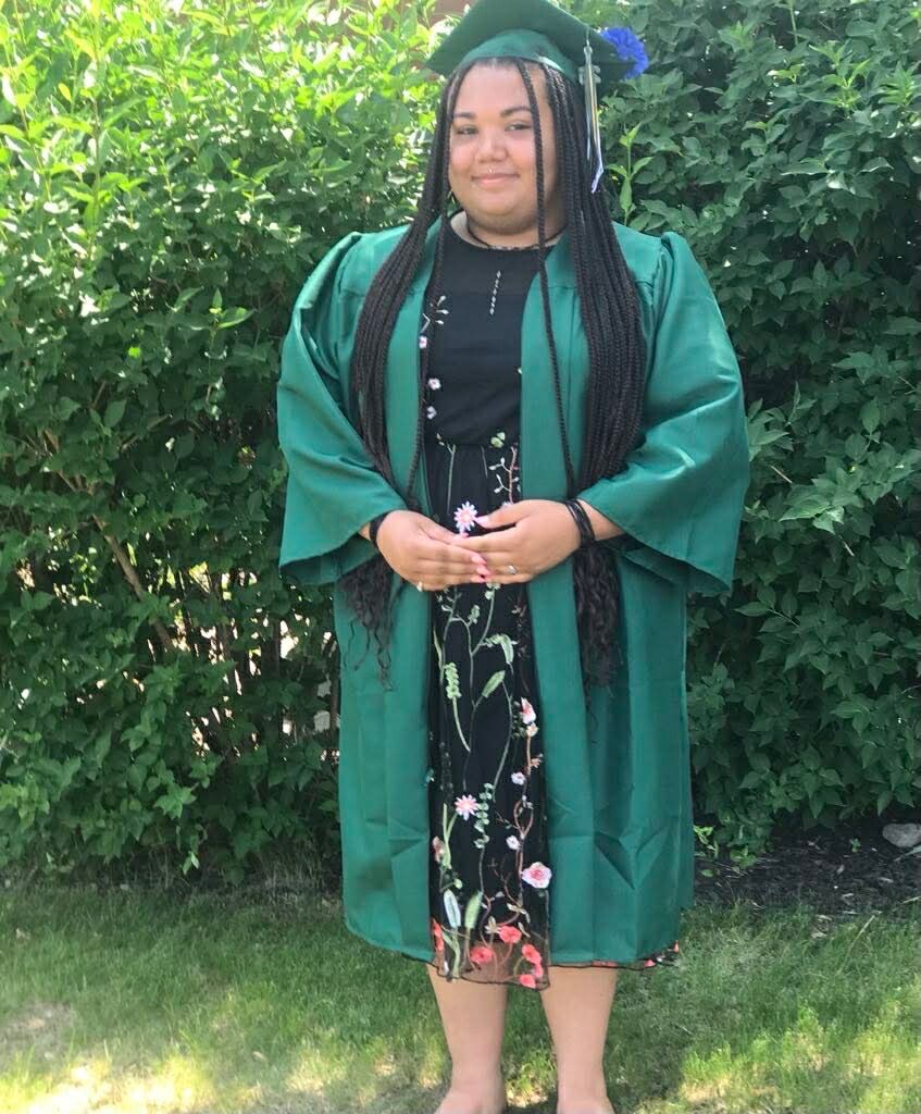 Graduate wearing a green cap and gown, standing outdoors in front of bushes, smiling, with a floral dress visible beneath the gown and cap tassel on her head.