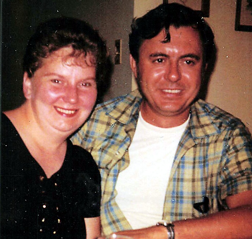 Two adults sitting together indoors, smiling at the camera; woman in black, man in a plaid shirt.