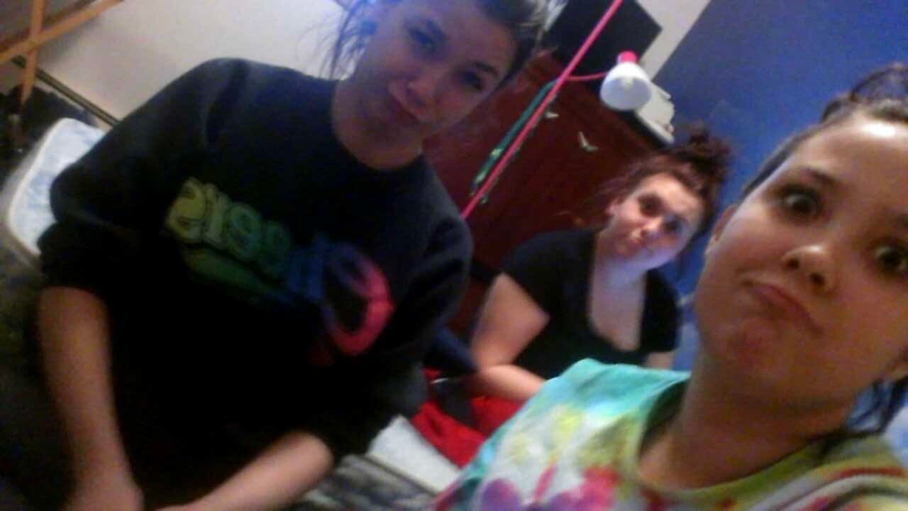 Three friends in a bedroom taking a group selfie; person in foreground wears a tie-dye shirt, two others sit in the background and smile at the camera.