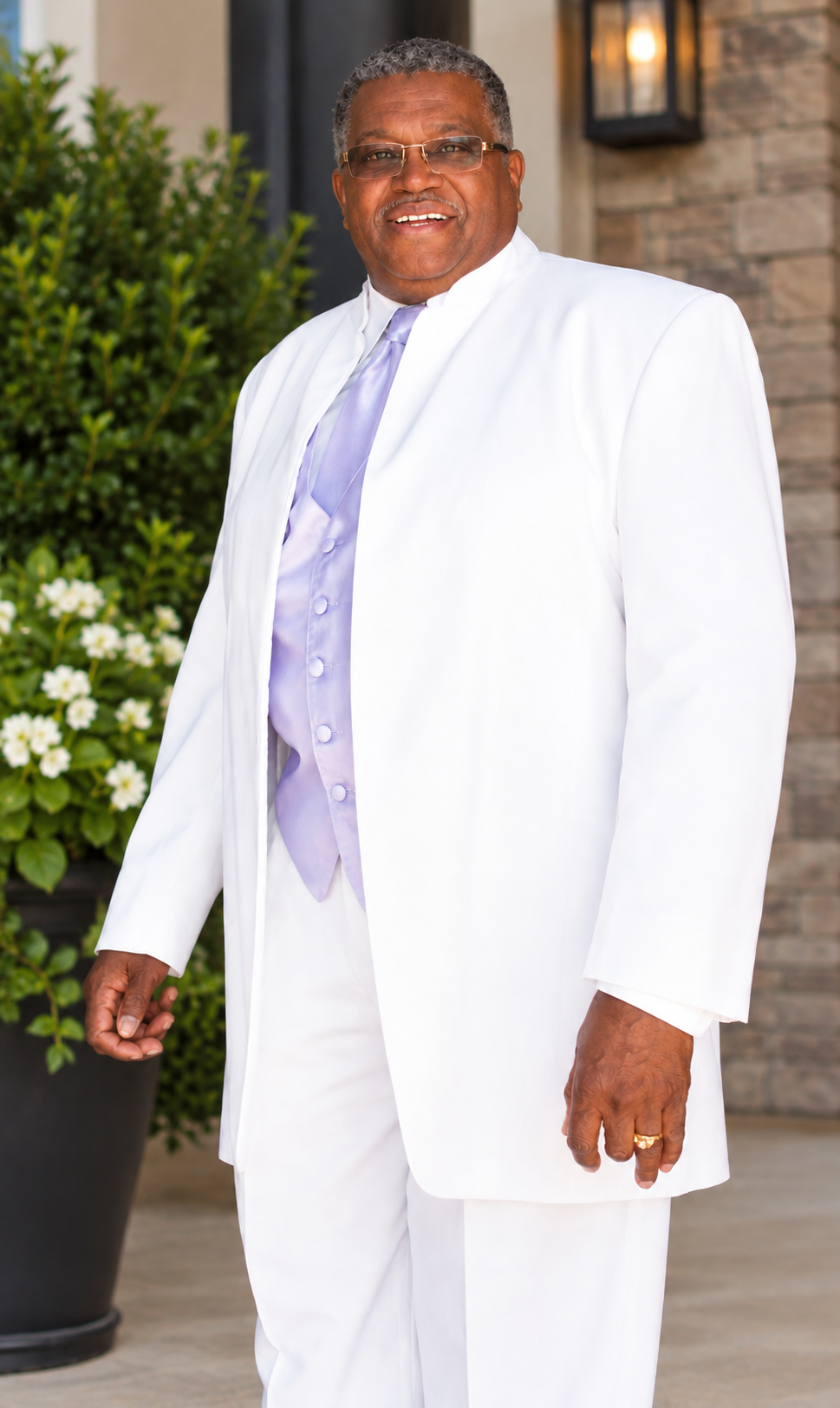Smiling man in a white suit with a lavender vest and tie, standing outdoors near greenery and a stone wall.