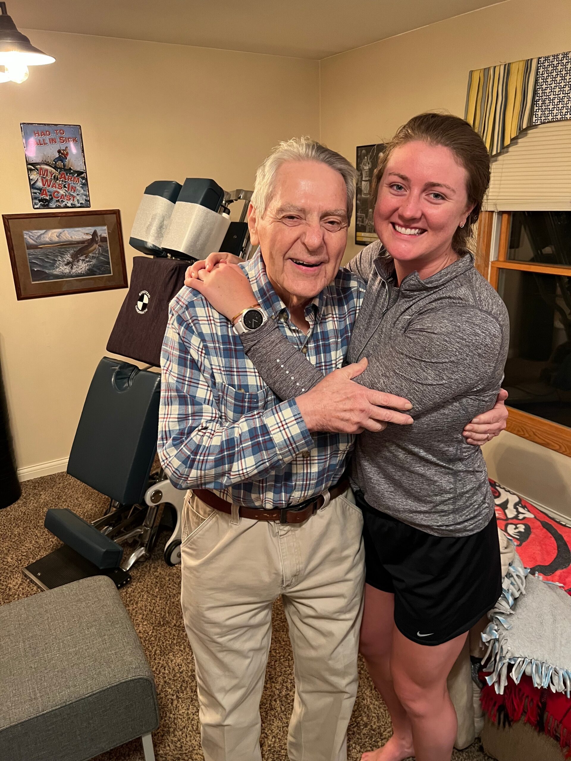 Two people hugging in a cozy room, an older man in a plaid shirt and a younger woman in gray, smiling at the camera.