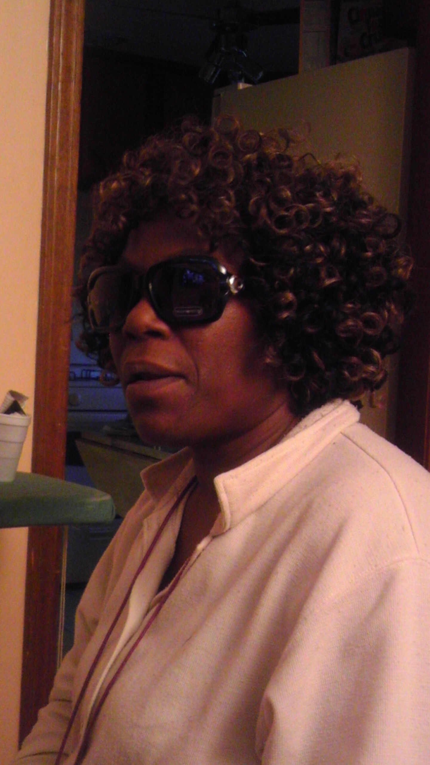 Woman with short curly hair wearing oversized sunglasses and a light-colored jacket, standing in a doorway indoors.