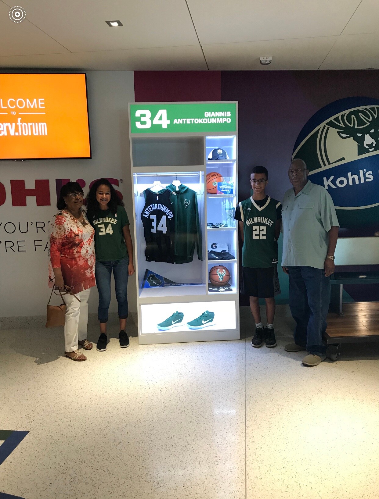 Four people stand next to a lit display case showcasing Milwaukee Bucks jerseys and a Giannis Antetokounmpo number 34 sign in a mall or store setting.
