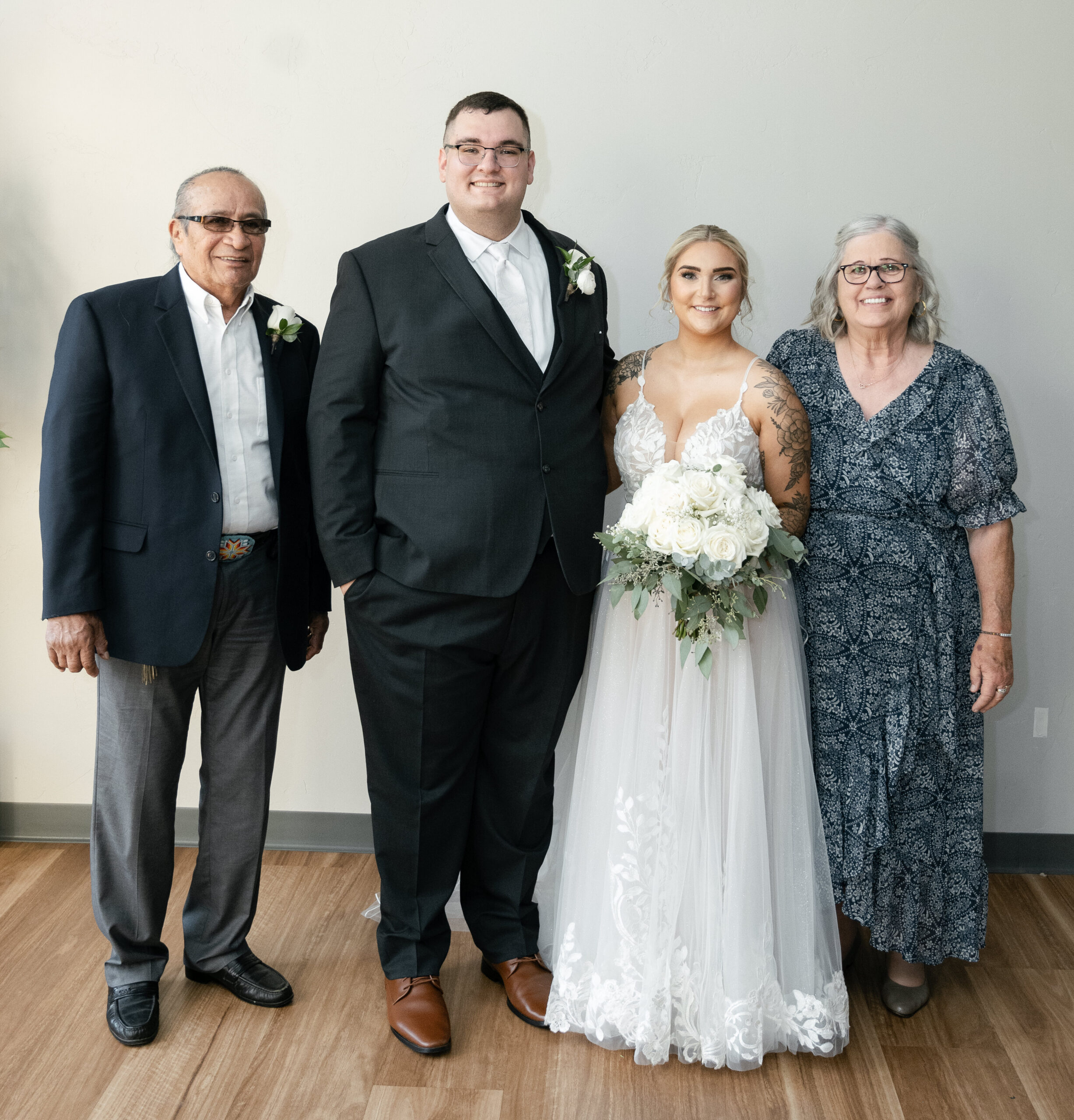 Group portrait of four adults indoors: groom in a black suit and white tie, bride in a lace white wedding gown holding a bouquet, an older man in a dark blazer with a boutonniere, and an older woman in a blue patterned dress posing together against a light wall.