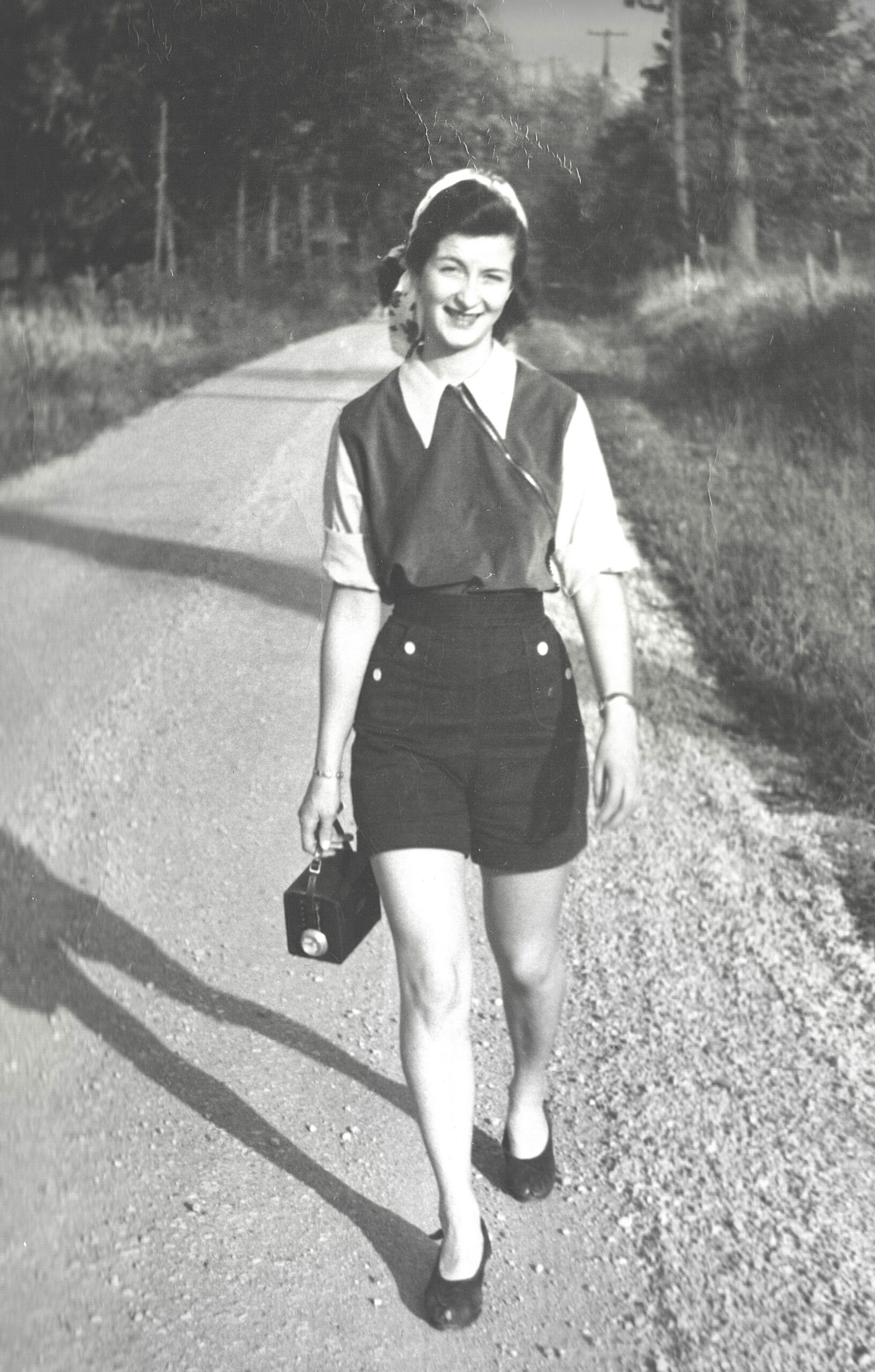 Young woman walking on a country road, smiling, carrying a vintage box camera and a small bag; wearing a blouse, vest, high-waisted shorts and flats.