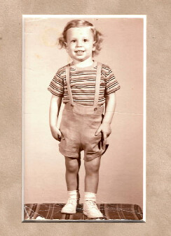 Young child standing and smiling, wearing a striped shirt with suspenders and short overalls, white socks and shoes, in a vintage sepia-toned photo.