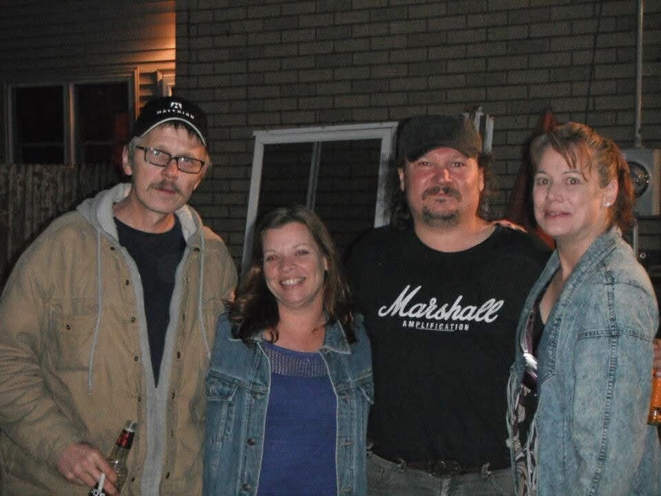 Four adults posing for a photo outside at night; man left in beige jacket holding a beer, two women in denim jackets, man in black Marshall t‑shirt in the middle.