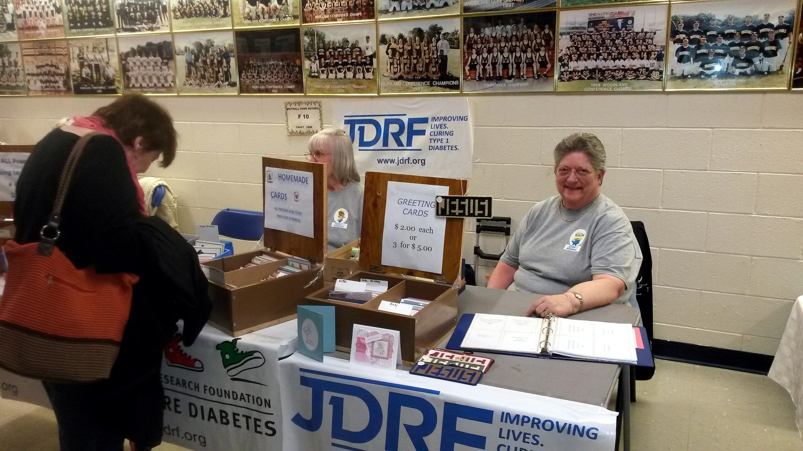 Booth at a community fair with a JDRF banner; a smiling volunteer sits behind a table selling homemade cards while a shopper browses nearby. Wall of group photos in the background.