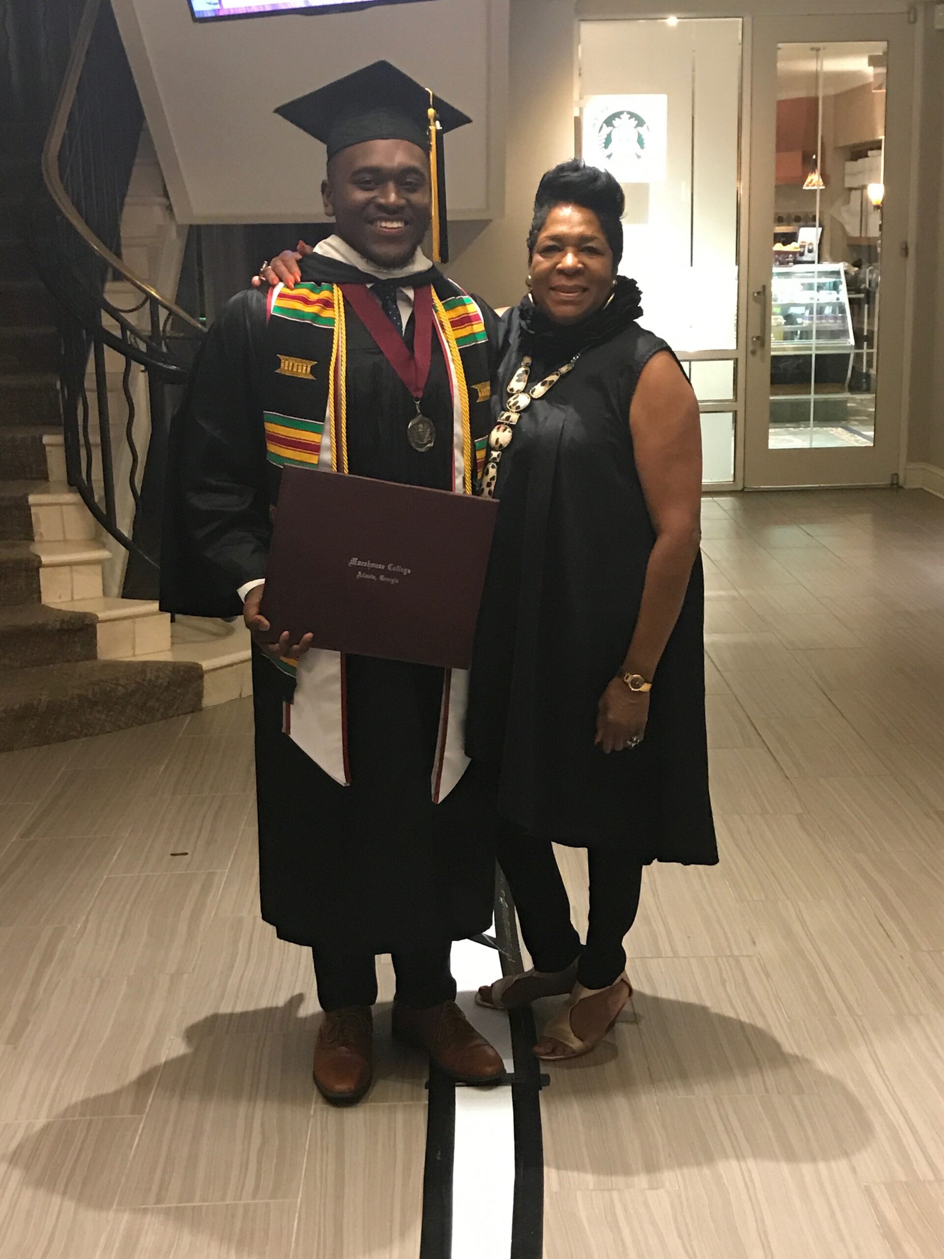 Graduate in cap and gown proudly posing with a woman, holding a diploma folder in a hallway interior.