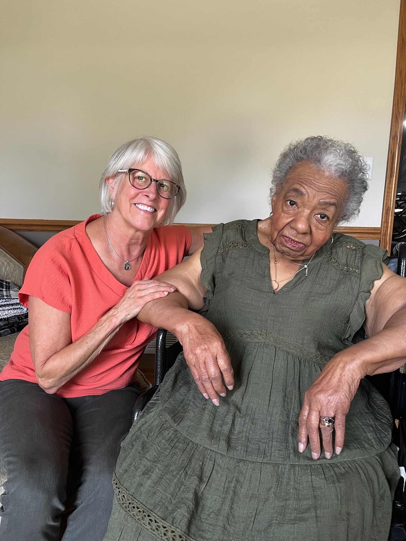 Two smiling women sit closely together indoors, one with white hair and glasses in a coral top, the other with gray hair in a green dress with a lace trim.