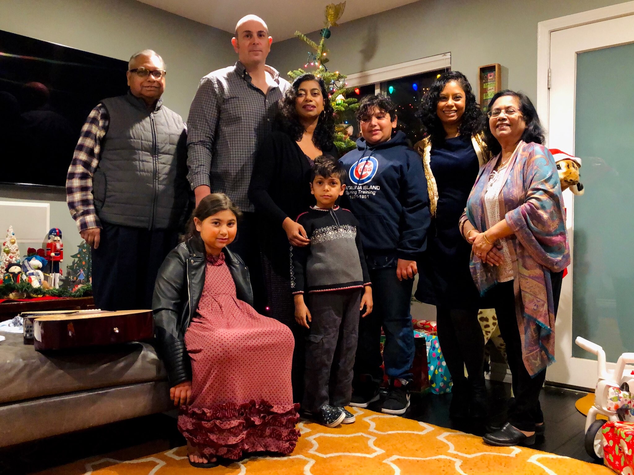 A diverse group of adults and children posing together for a holiday photo in a festively decorated living room, with a small Christmas tree and wrapped gifts nearby.