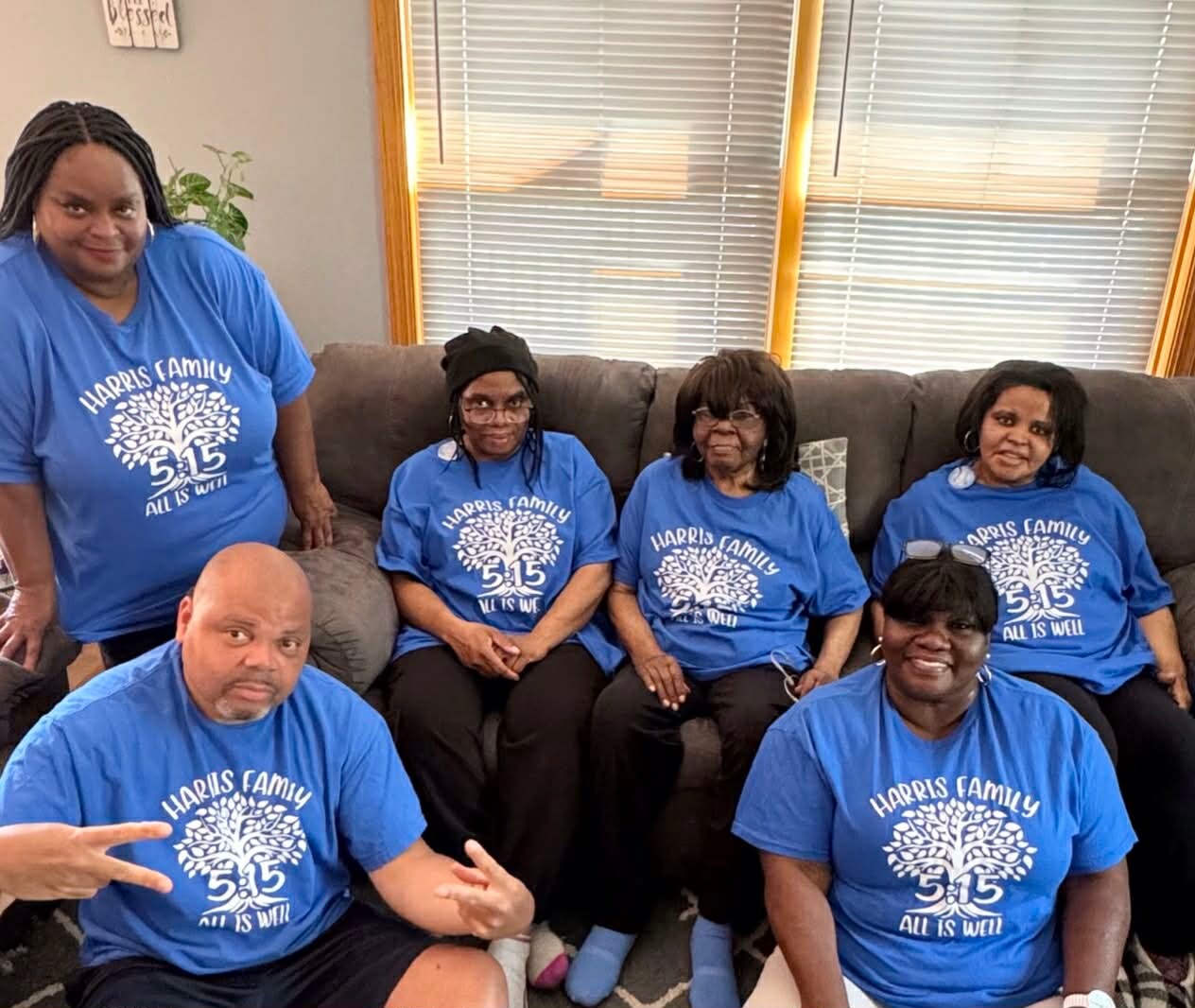 Six adults wearing matching blue Harris Family shirts sit and pose on a living-room couch, smiling at the camera.