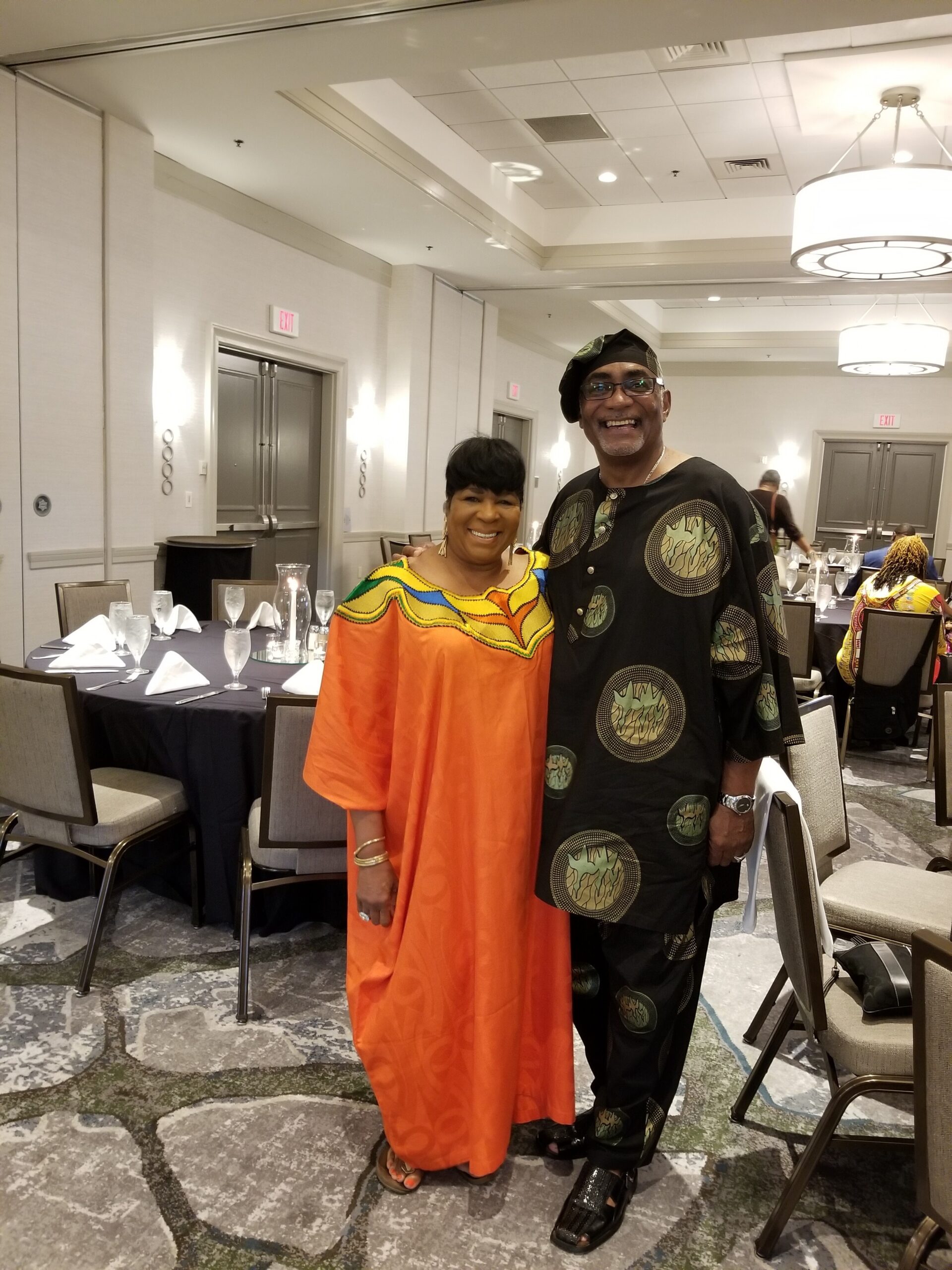 Smiling couple in a banquet hall wearing vibrant traditional outfits; woman in orange dress, man in black patterned attire.