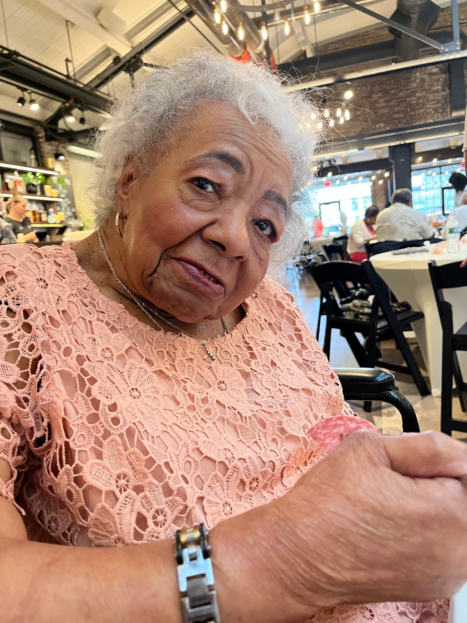 Close-up of an elderly woman with white hair wearing a pink lace top, smiling in a bright cafe setting.