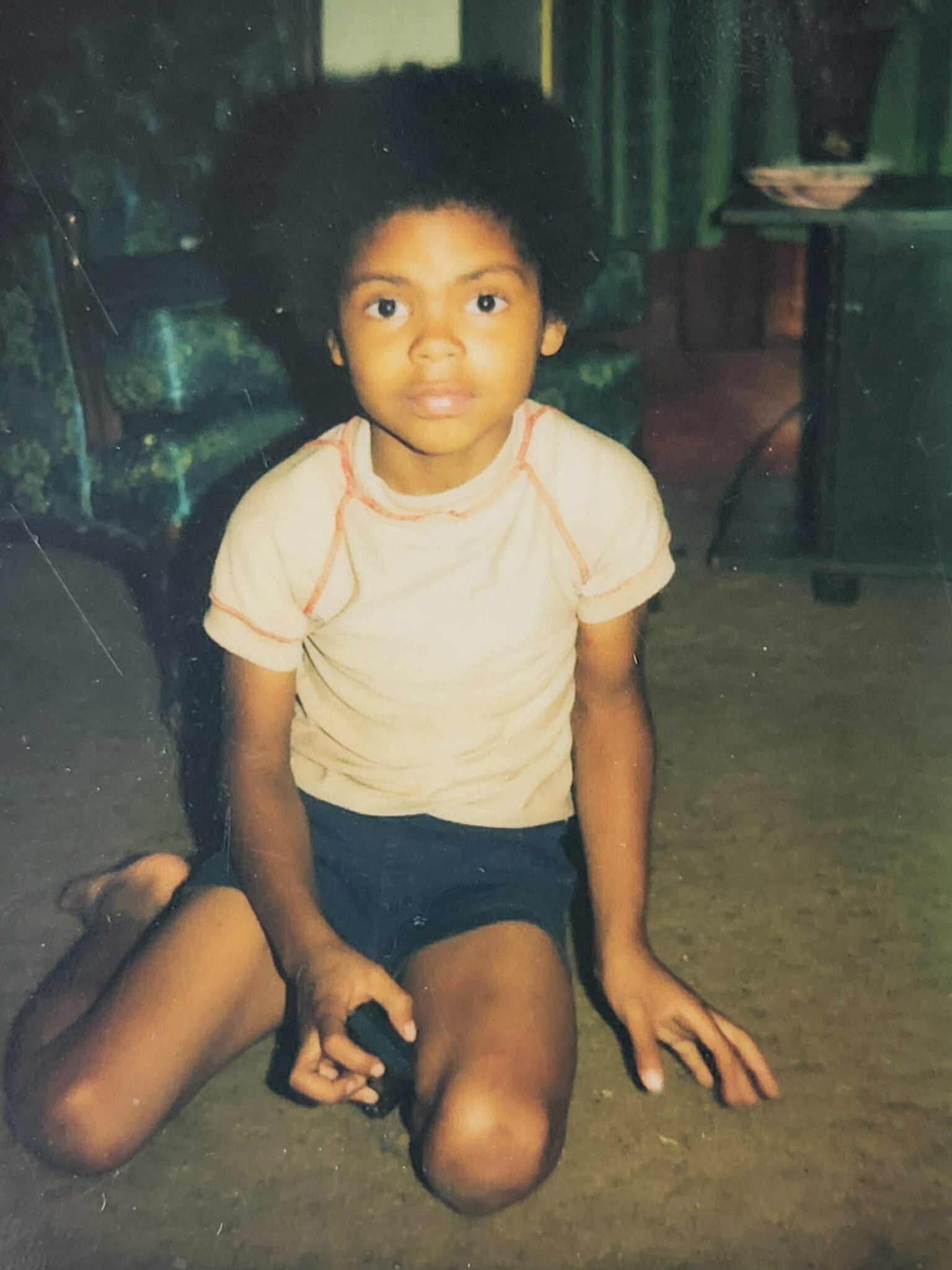 Young child with curly hair kneeling on a dusty floor, wearing a light shirt and dark shorts, looking at the camera.