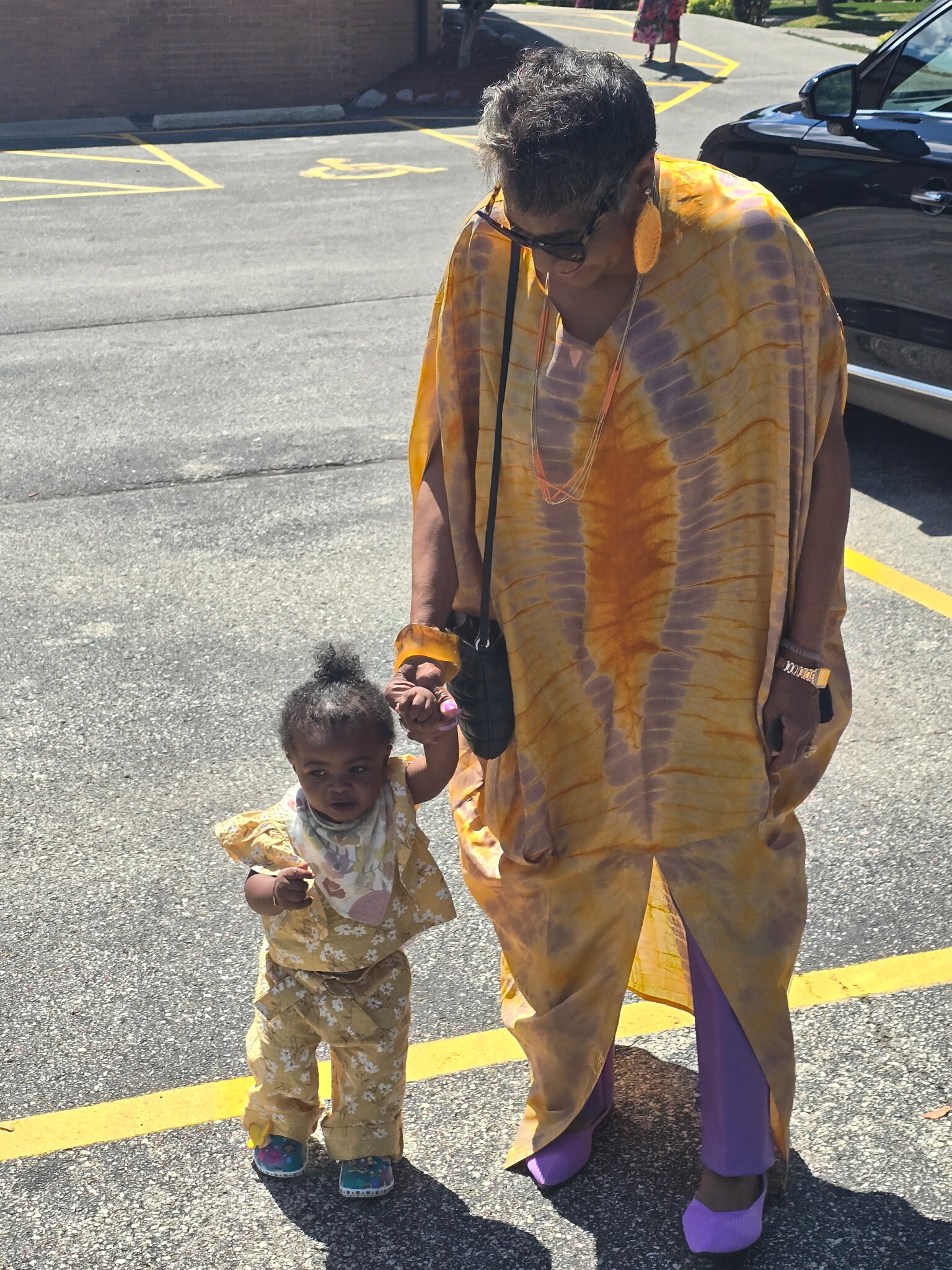 Adult in a yellow tie-dye outfit holding hands with a toddler in matching clothes in a parking lot.
