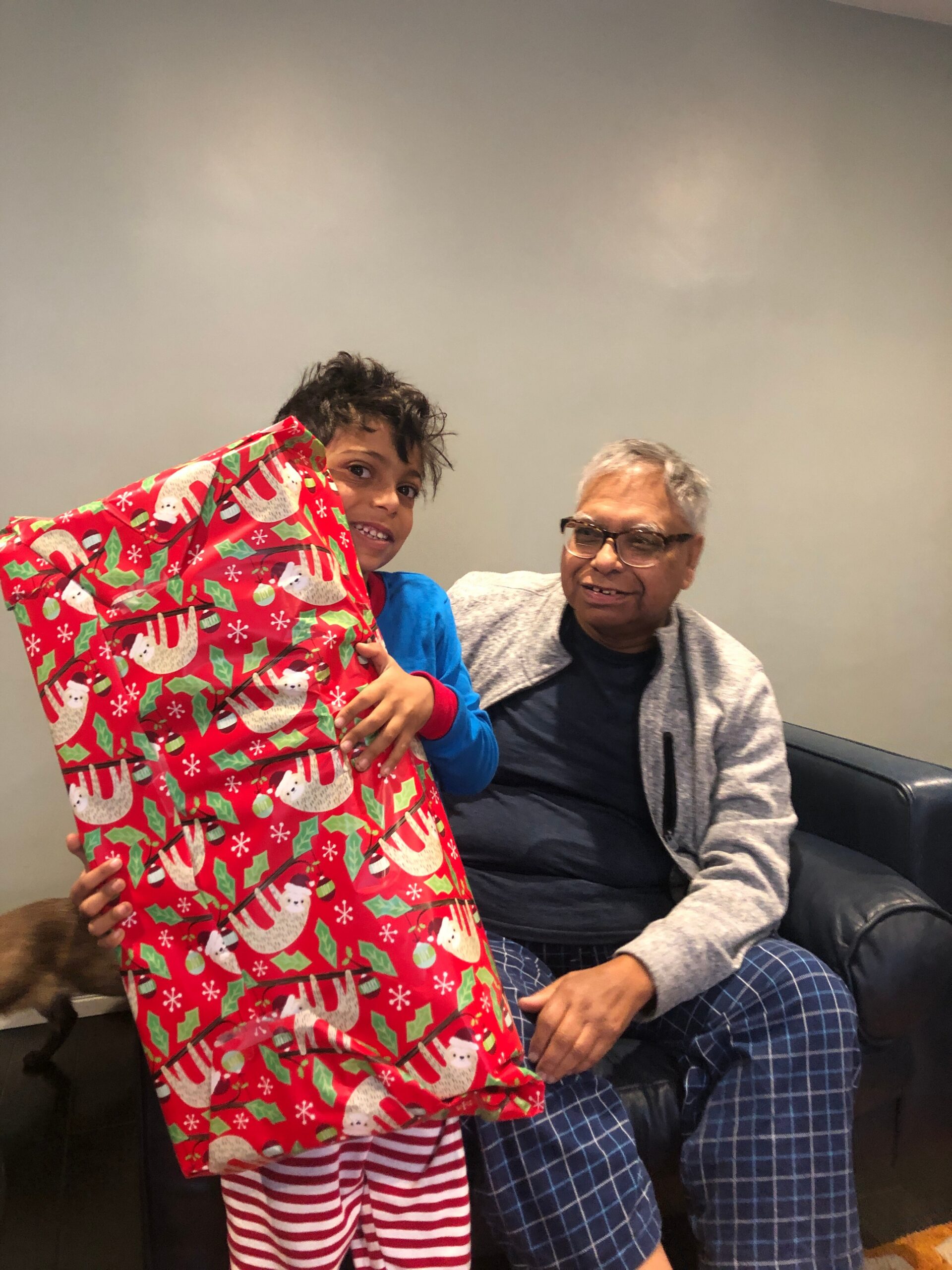 Two people near a black couch: a smiling boy holding a large red Christmas gift, while an older man sits beside him.