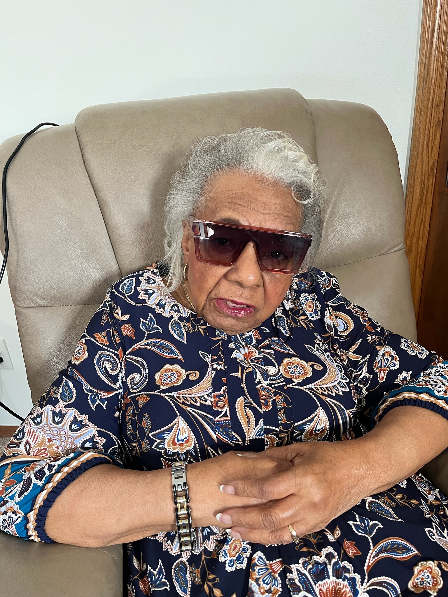 Older woman with short gray hair wearing red sunglasses, seated on a beige chair, hands folded on lap in a patterned navy blouse.