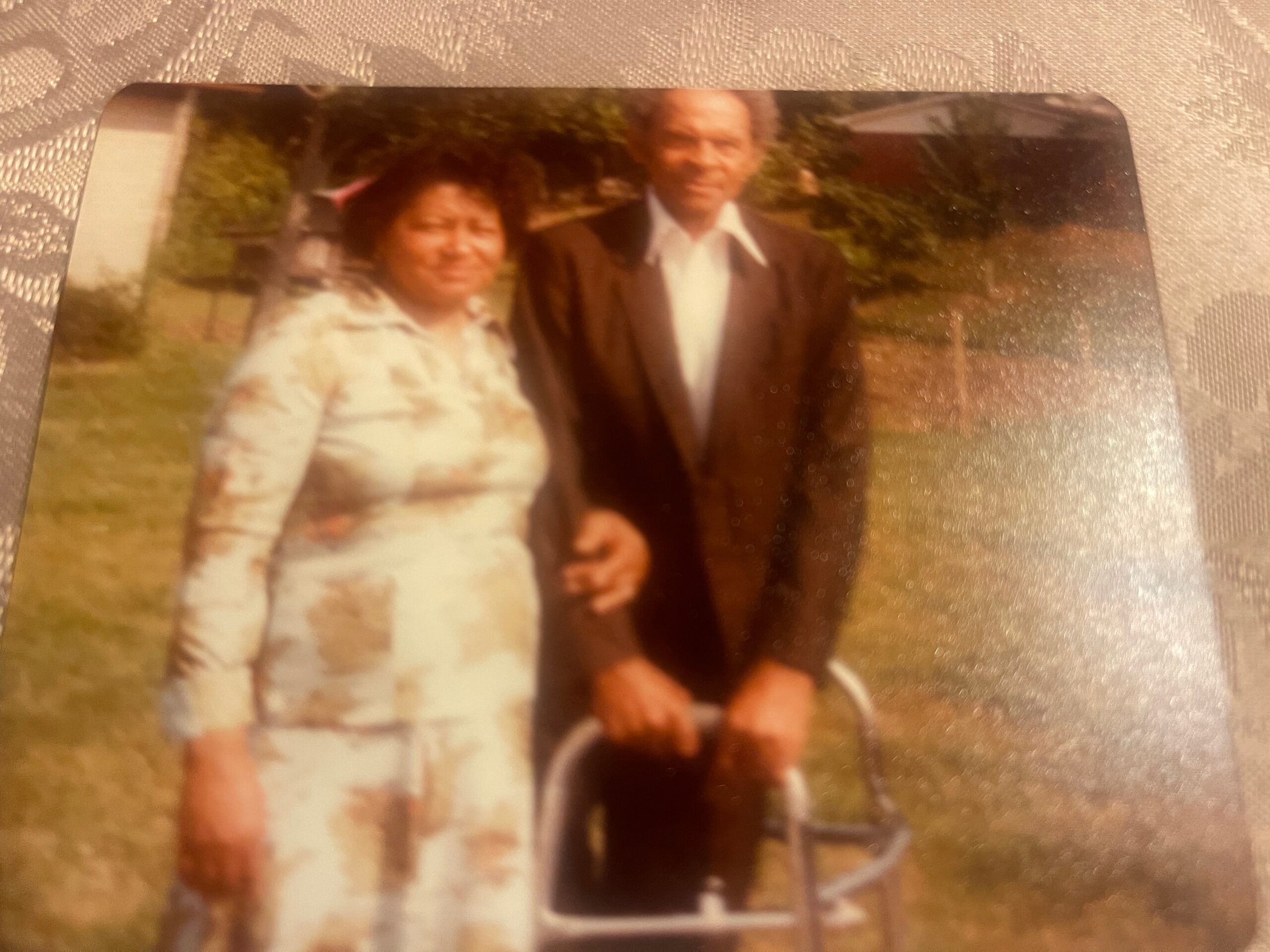 Two people posing outdoors: a woman in a light dress and a man in a dark blazer, with greenery in the background (blurry).