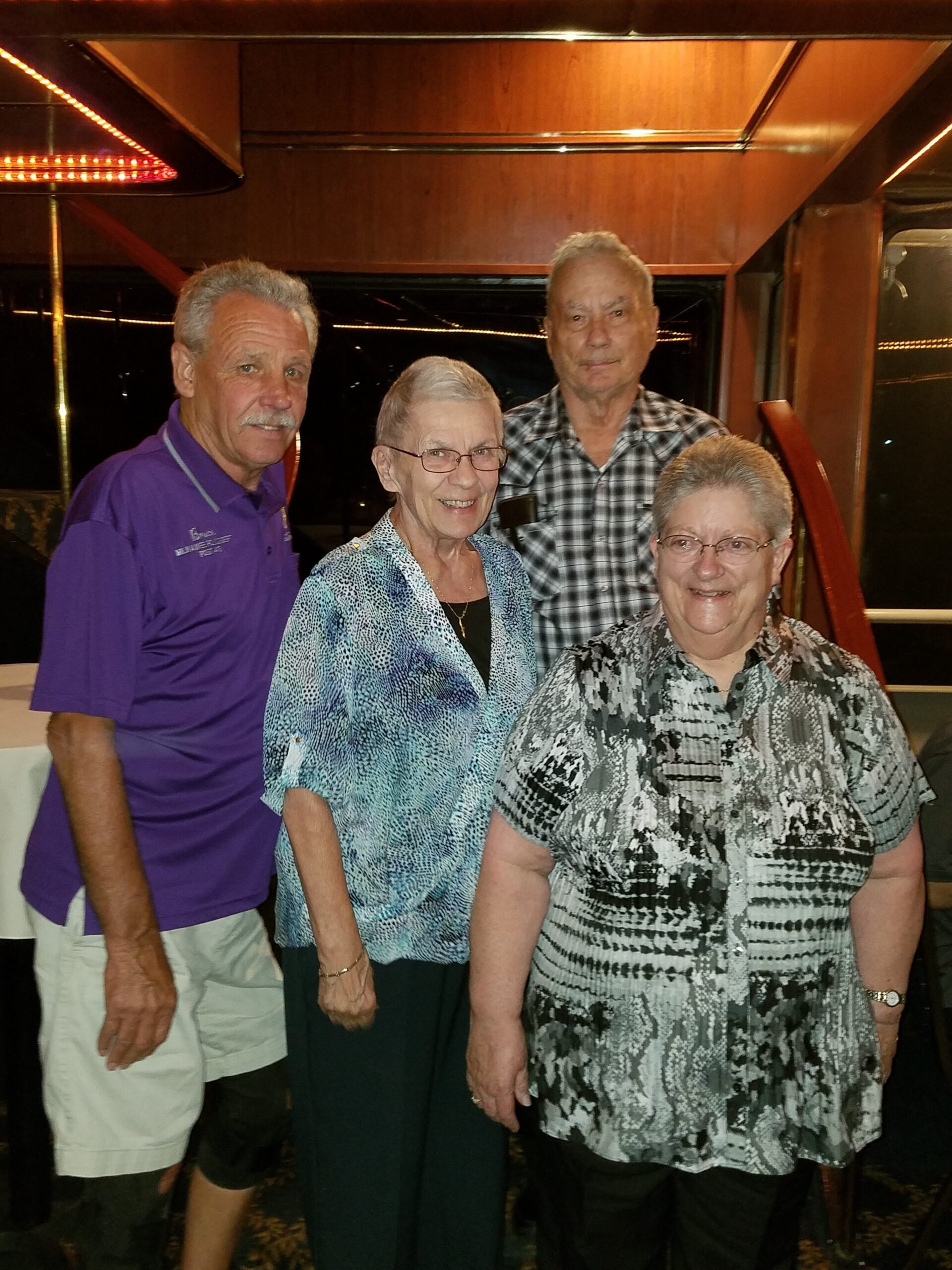 Four smiling elderly people posing together indoors, two men and two women, dressed casually.