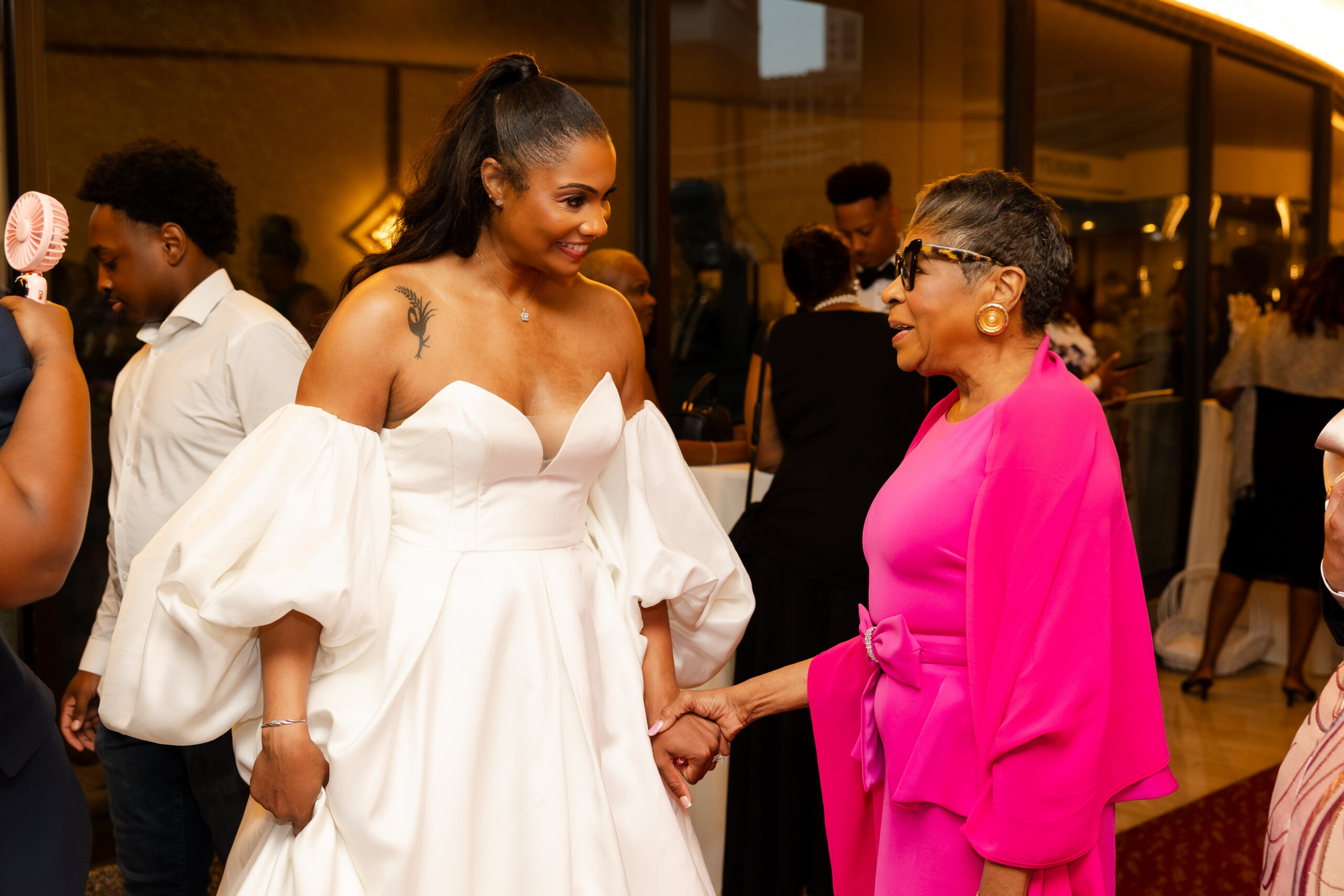 Woman in a white off-shoulder gown smiles while holding hands with an older woman in a bright pink outfit at a formal event.