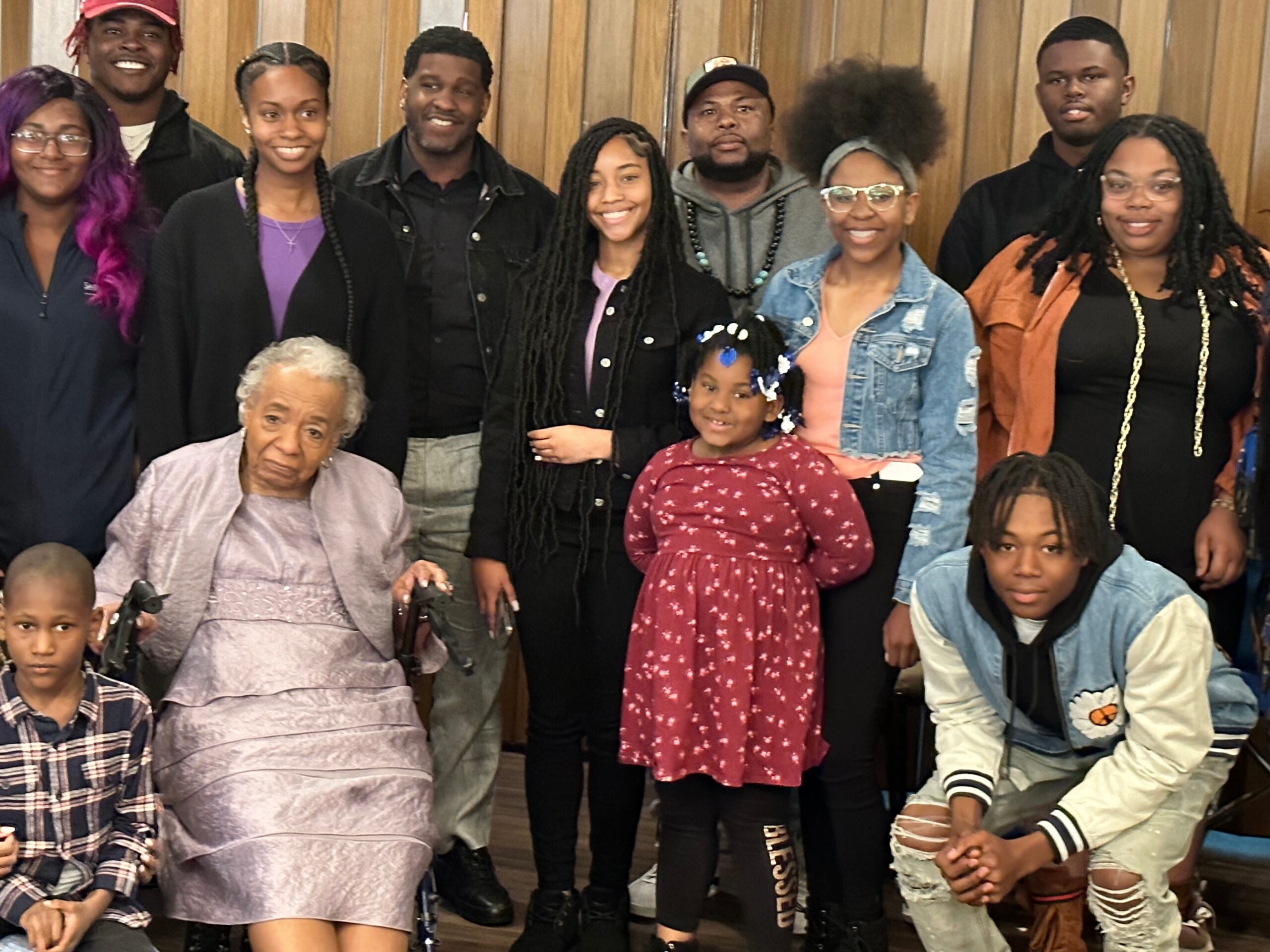 A diverse group of people, including an elderly woman in a purple dress, posing together and smiling indoors.