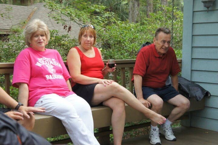 Three adults sit on a wooden deck railing outdoors; the woman on the left wears a pink shirt and white pants, the woman in the middle holds a glass of red wine, and the man on the right wears a red polo and shorts.
