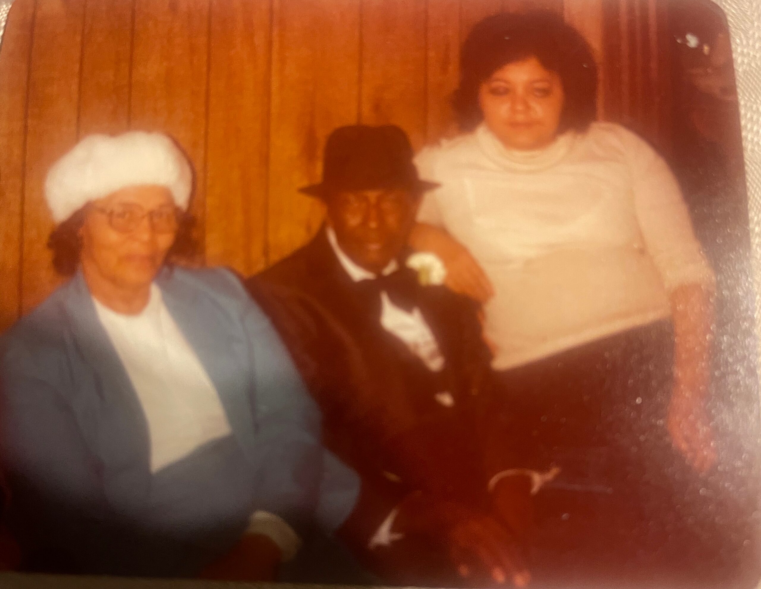 Three people posing for a group photo indoors with wood-paneled walls: left, an older woman with white hair and glasses; center, a man in a dark suit and hat; right, a woman in a light-colored top.