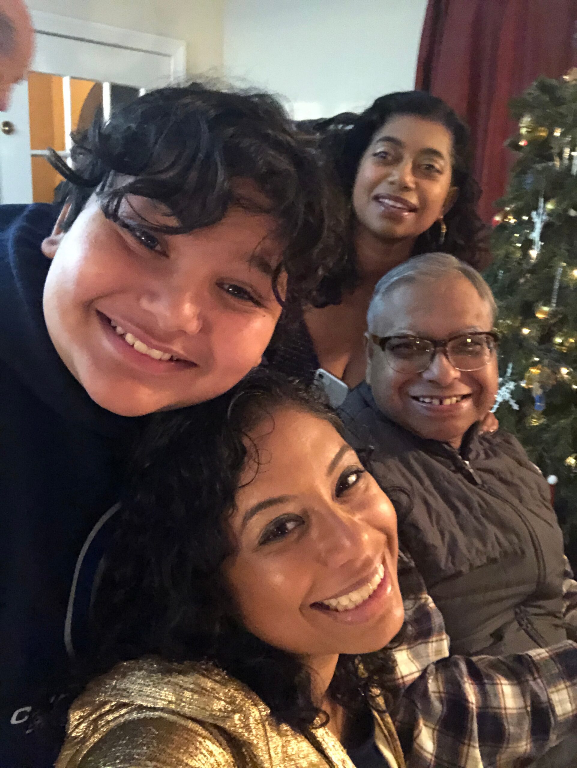 Four adults smiling for a close-up group selfie indoors, with a Christmas tree in the background.