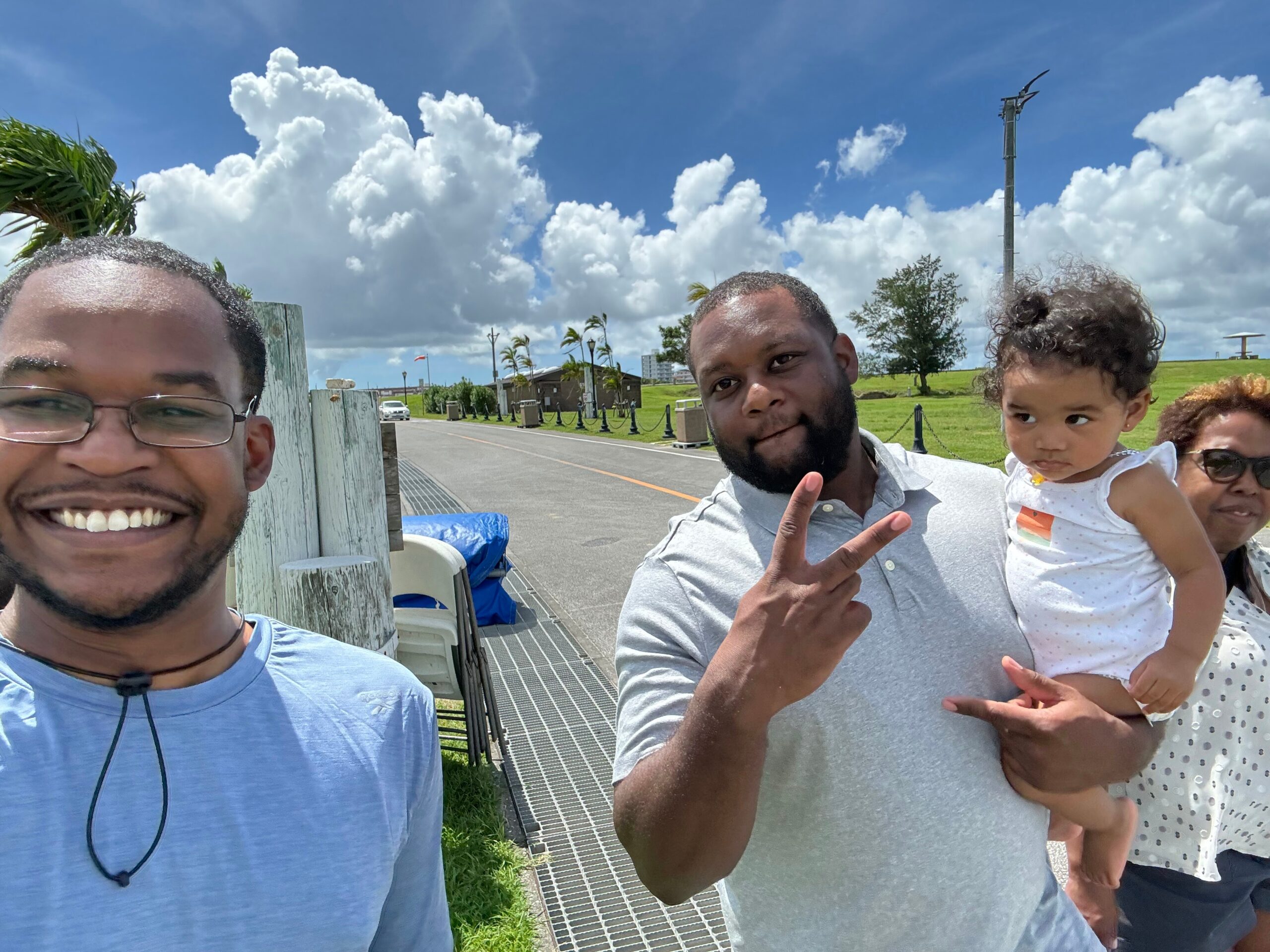 Smiling group selfie outside on a sunny day, with a man in a blue shirt, another man making a peace sign, and a child being carried against a bright sky and clouds.