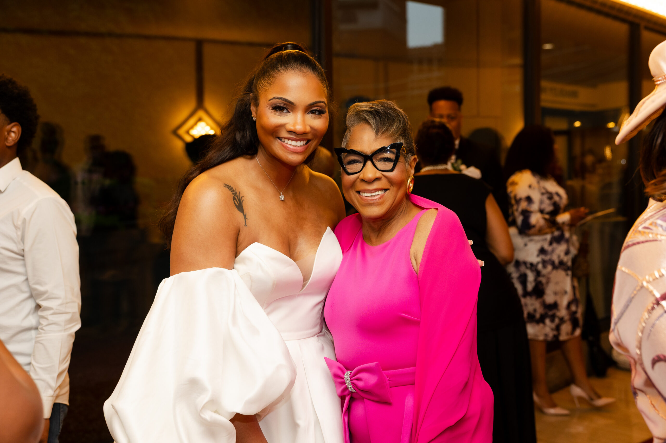 Two women posing together at a party, one in a white off-shoulder dress and the other in a bright pink outfit, smiling at the camera