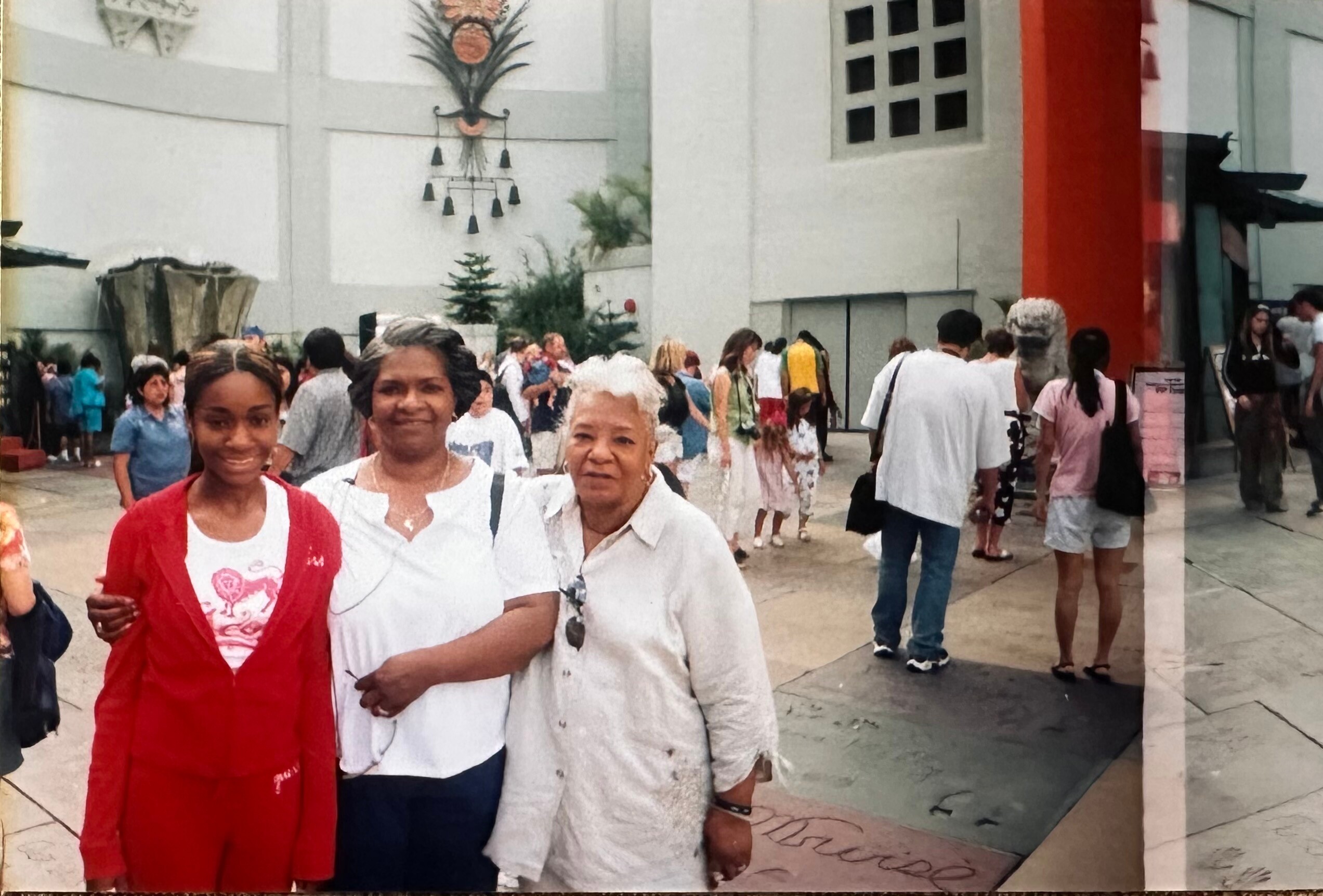 Two women in the foreground smile at the camera as a crowd gathers in a busy outdoor plaza with modern buildings behind them.