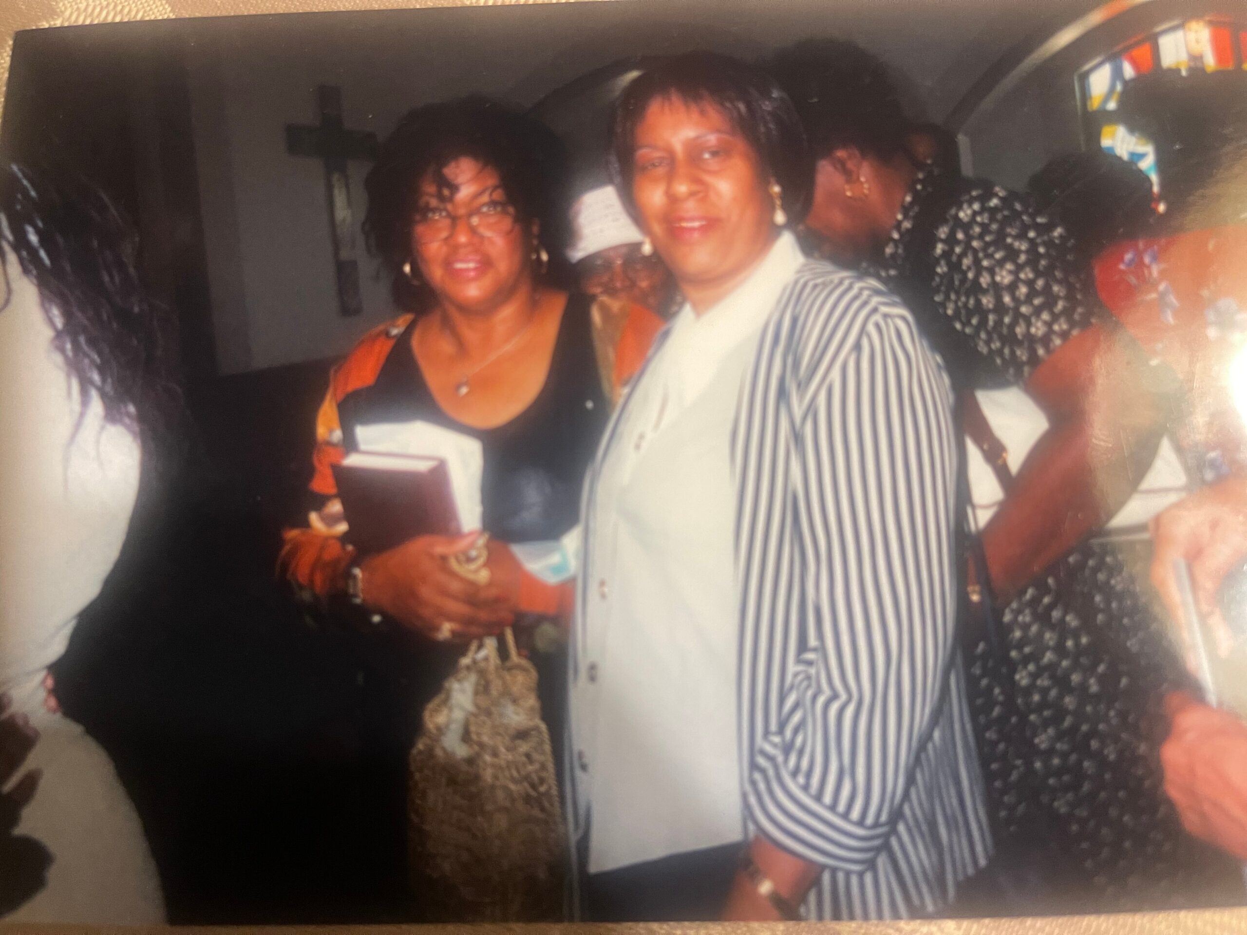 Two women smile for a casual group photo at a party; foreground woman wears a striped dress, another in black and orange attire beside her.