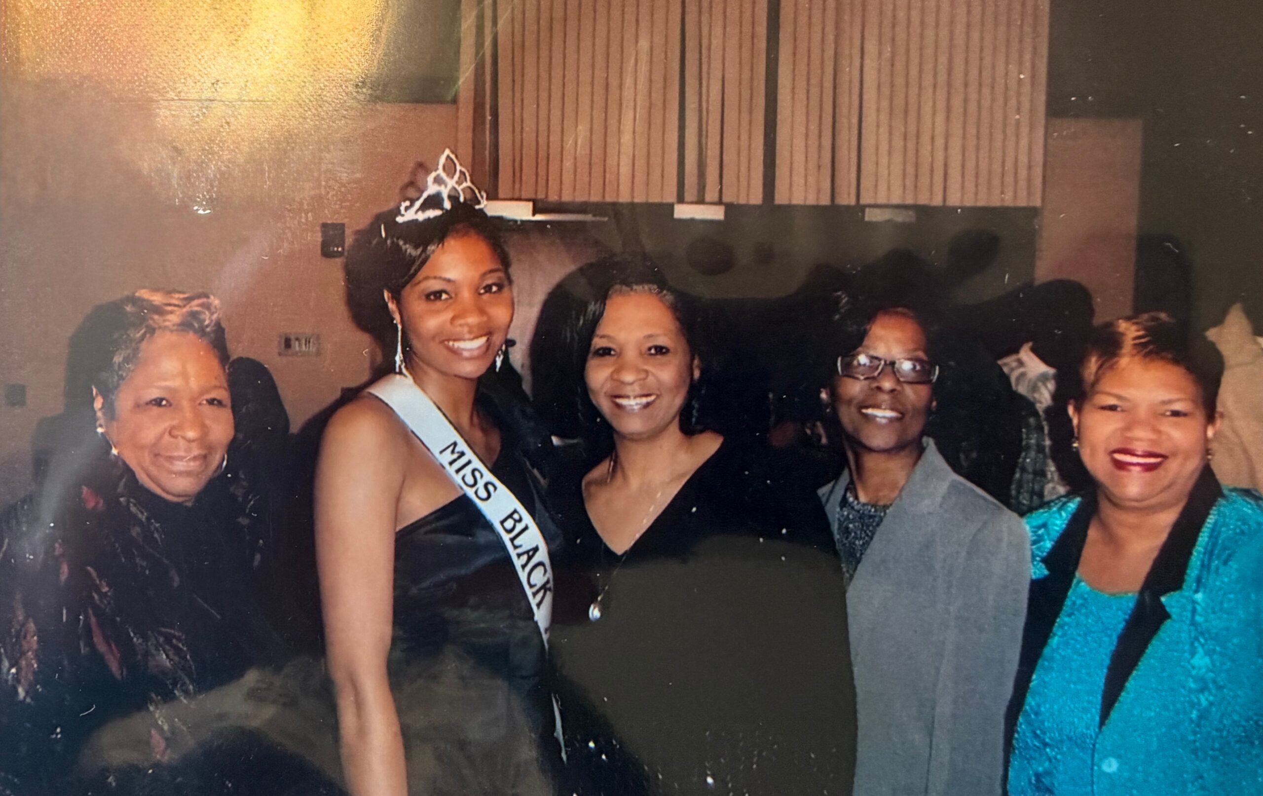 Smiling group of women at a social event; the central woman wears a tiara and a 'Miss Black' sash.