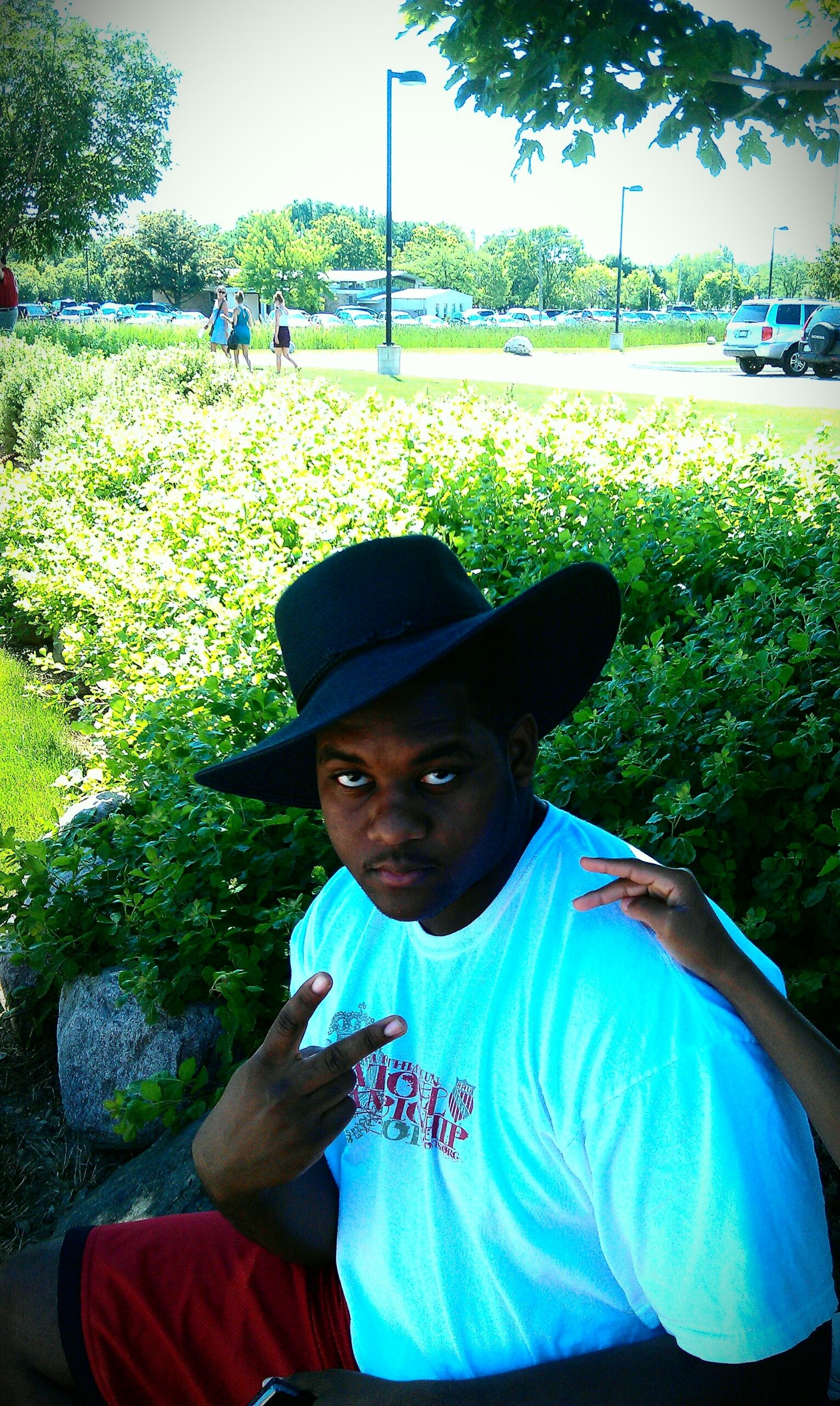 Young man in a black wide-brim hat and white T‑shirt, making peace signs in a sunny park with greenery and parked cars in the background.