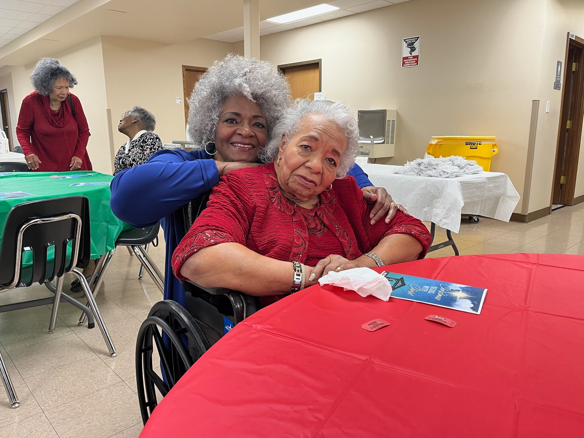 Two elderly women sharing a hug at a community event, seated by a red table with a wheelchair in the foreground.