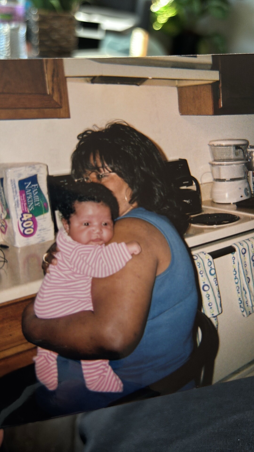Woman in a blue sleeveless top holds a baby wearing a pink striped onesie in a kitchen by the counter and appliances nearby.