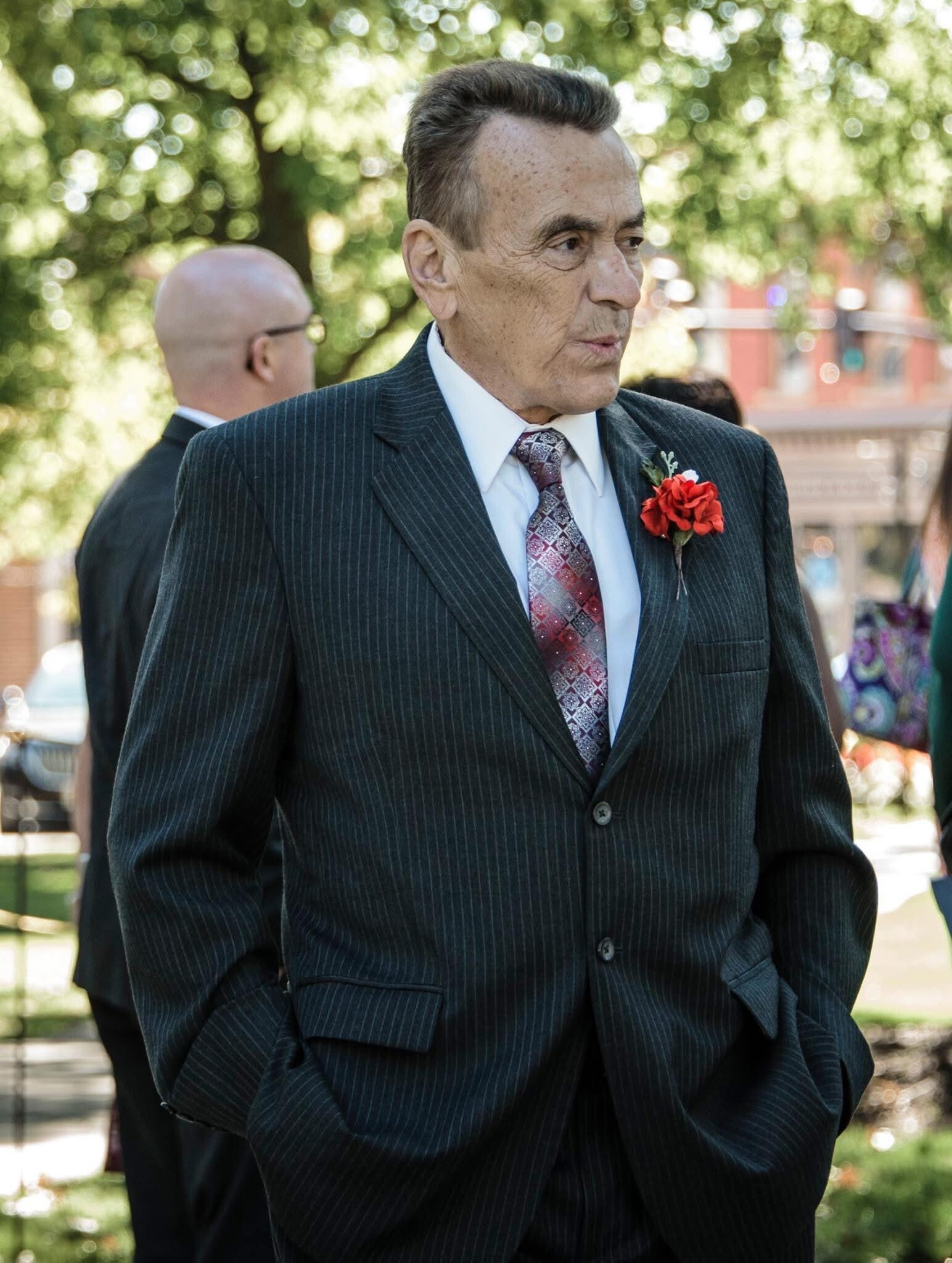 Older man in a dark pinstripe suit with a red carnation boutonniere, standing outdoors.