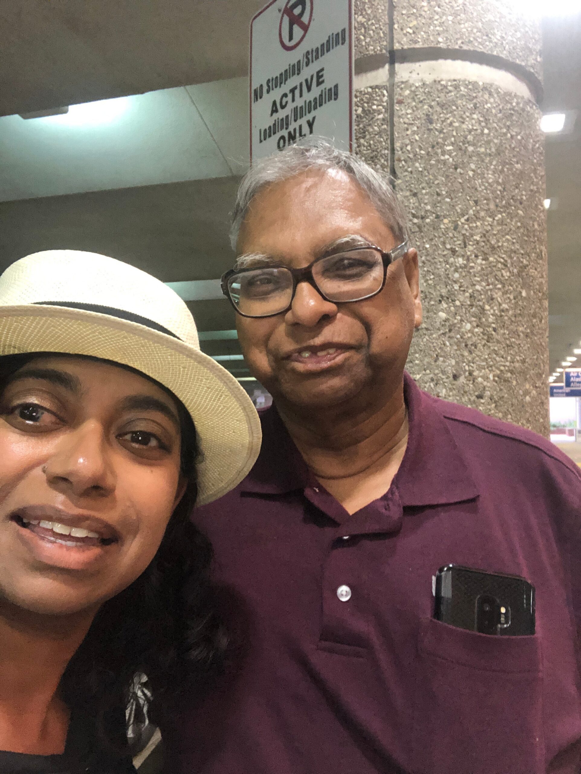 Two people take a selfie in a parking garage: an older man with glasses in a maroon polo smiles, while a woman wearing a wide-brimmed hat is partially visible in the foreground.