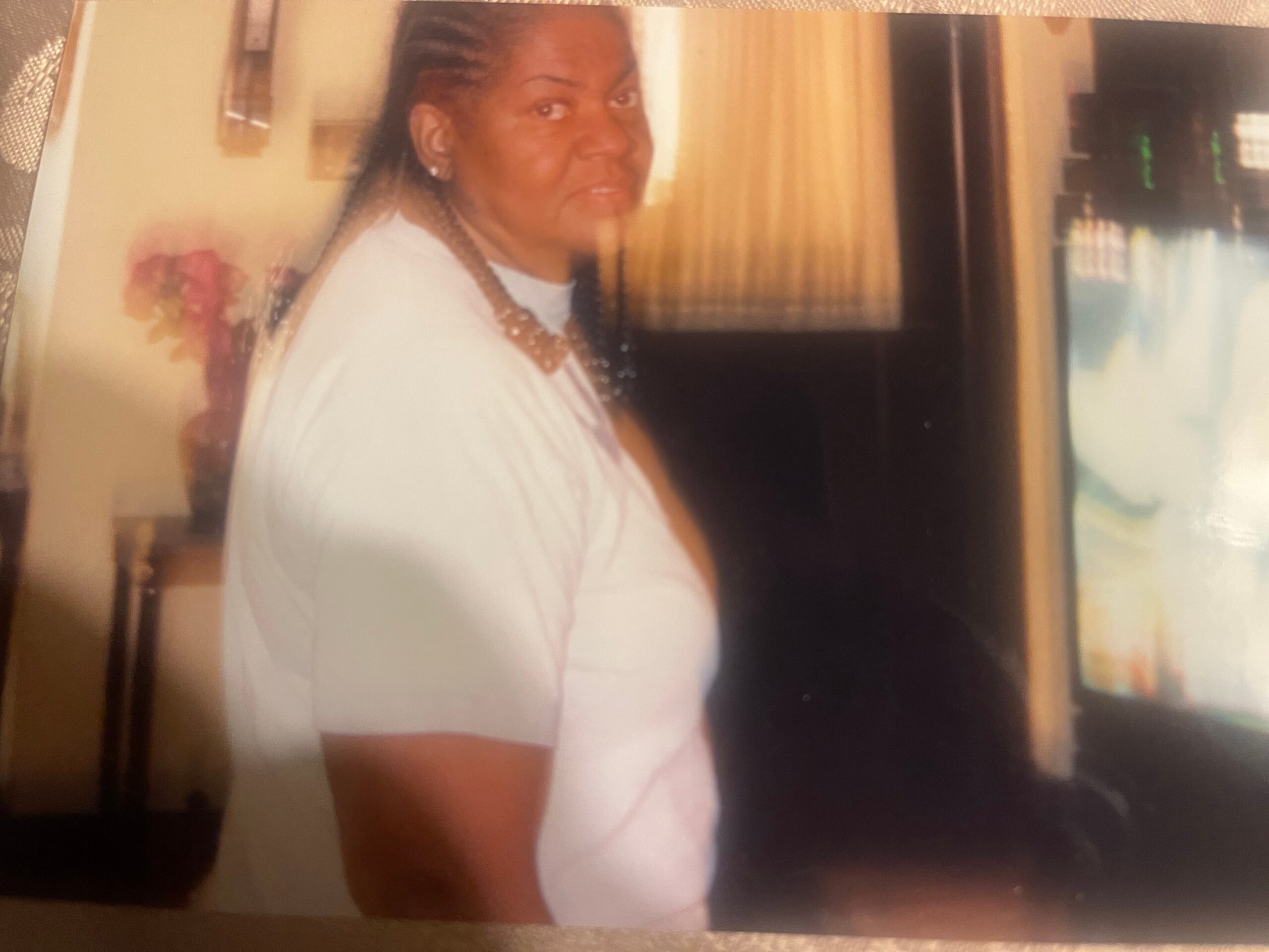 Woman with braided hair wearing a white shirt, standing in a kitchen near a refrigerator, looking toward the camera.