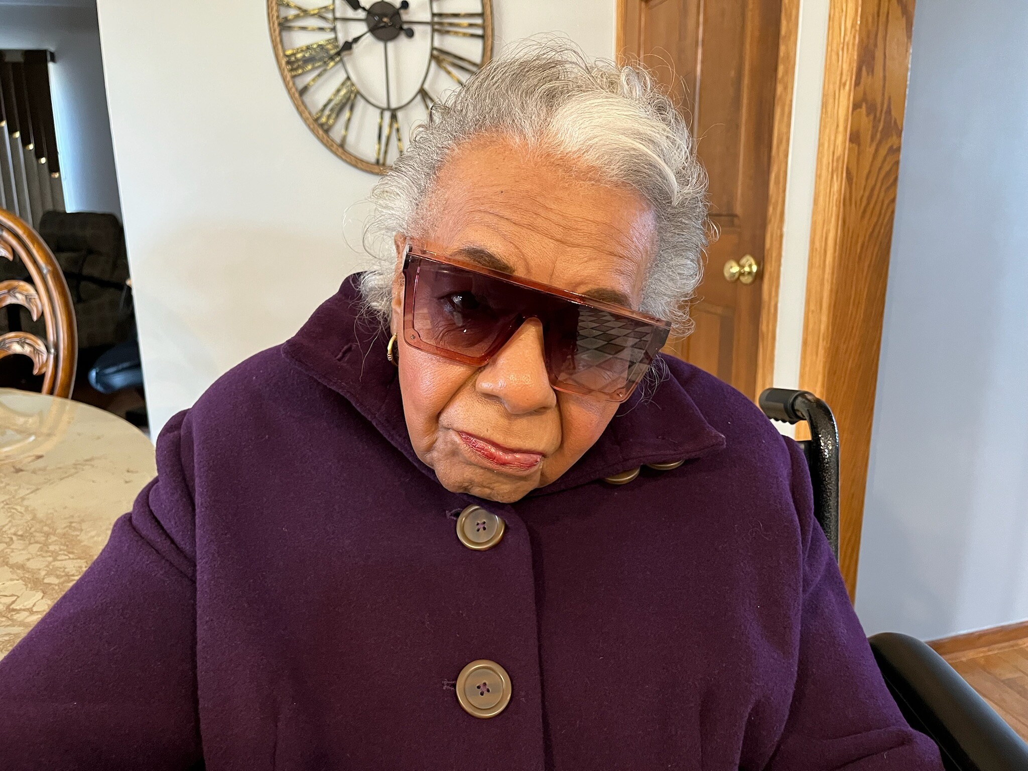Close-up of an older woman wearing oversized maroon sunglasses and a purple coat indoors, with a large clock on the wall behind her.