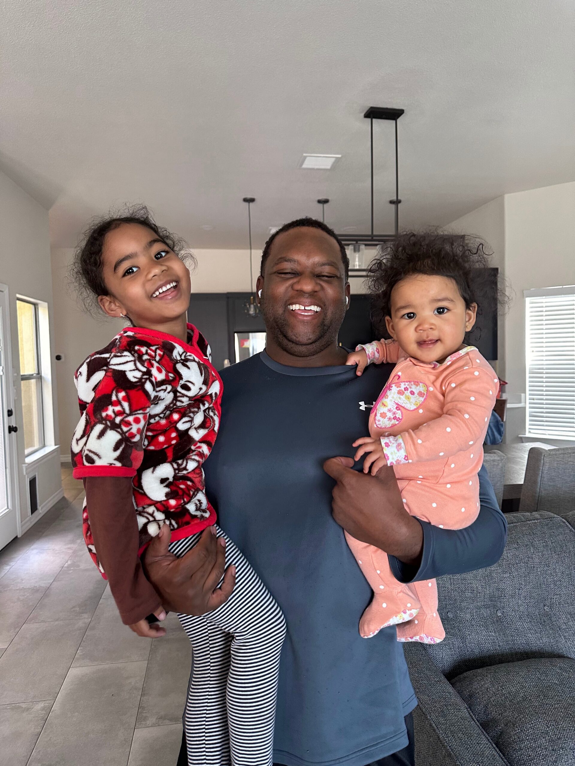 Father holding two young girls, all smiling inside a bright home living area with a gray couch in the background.